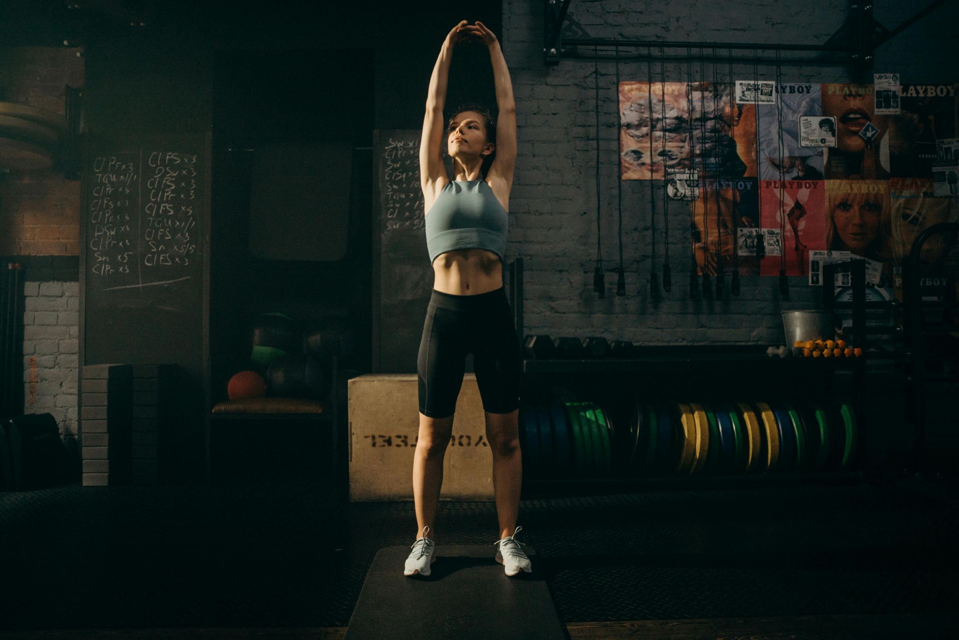Woman stretches arms overhead in a gym. She wears athletic clothing in front of weights and posters.