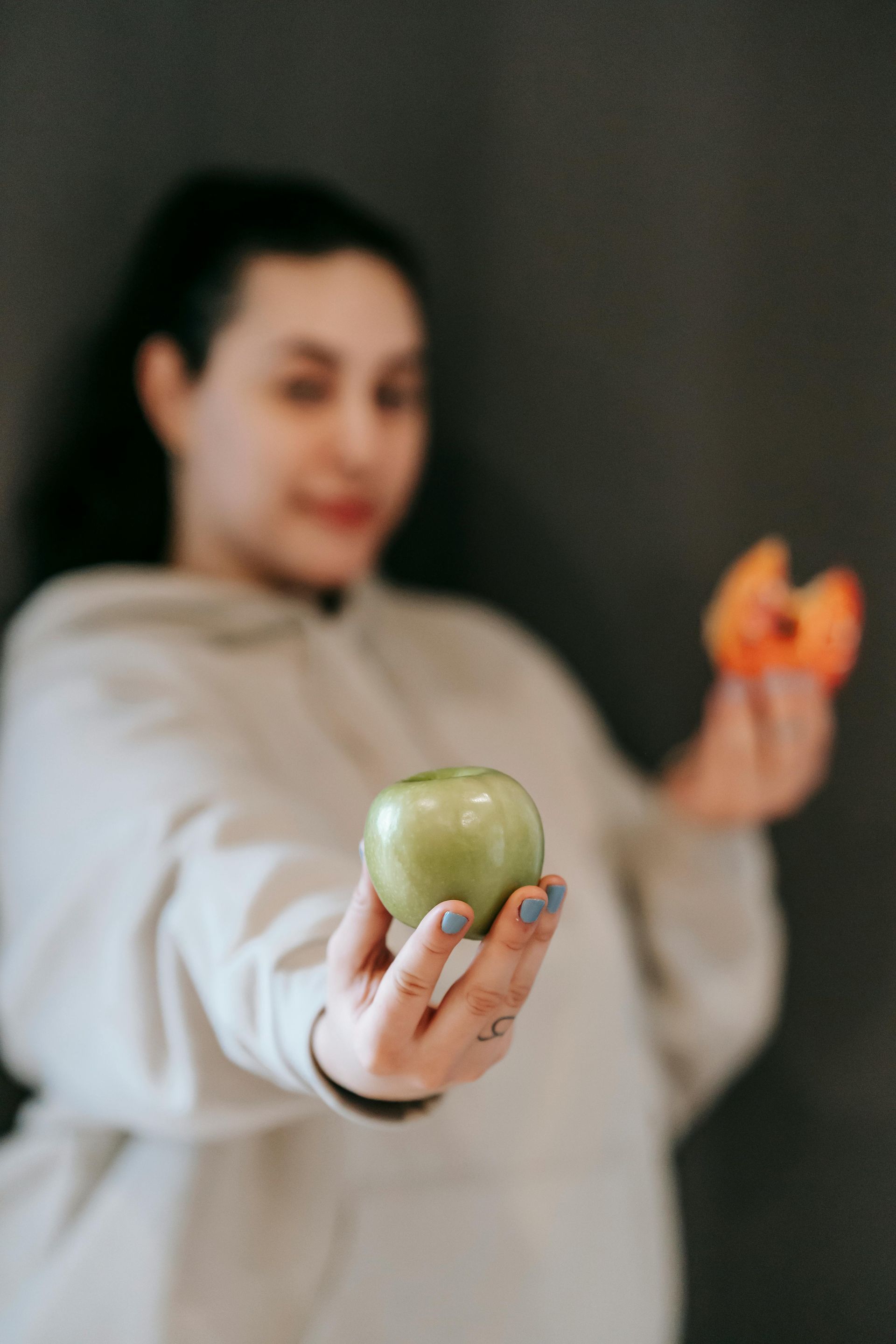 Woman holding a green apple and a bitten peach against a dark backdrop.