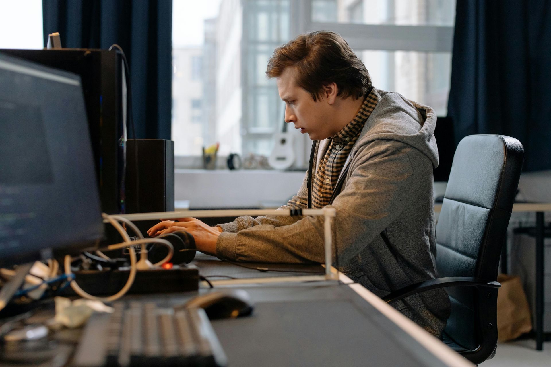 Man in a gray hoodie at a desk, focused on a computer. Office setting, window in the background.