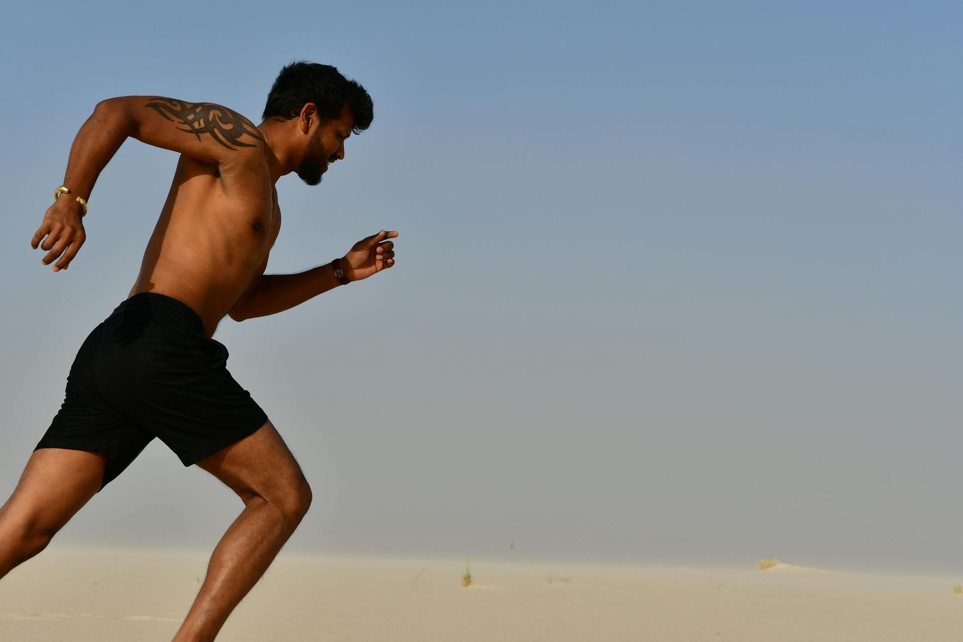 Man running on sand under a clear blue sky, wearing black shorts, with a tattoo on his arm.