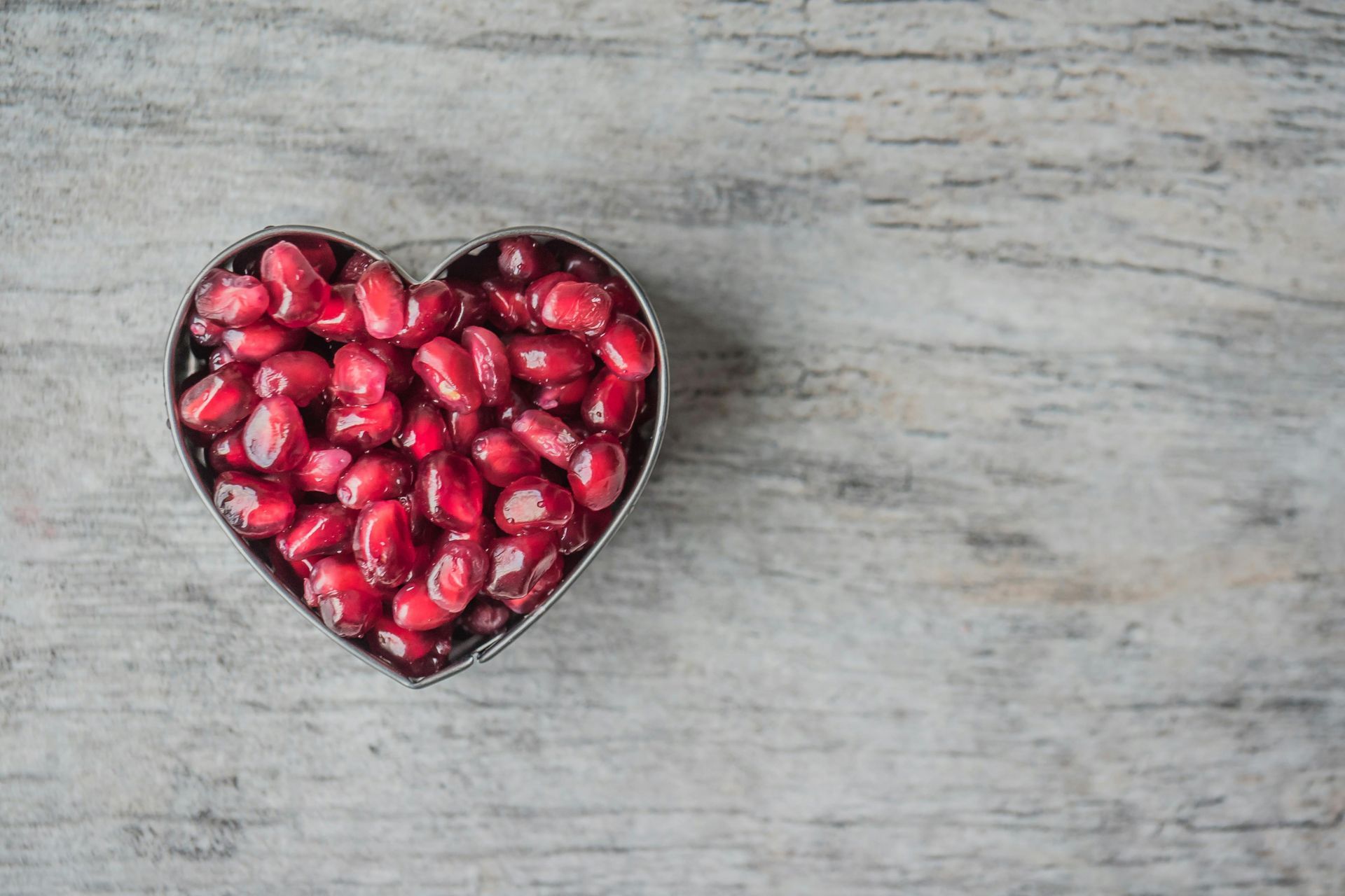 Heart-shaped dish filled with bright red pomegranate seeds on a light, weathered wooden surface.