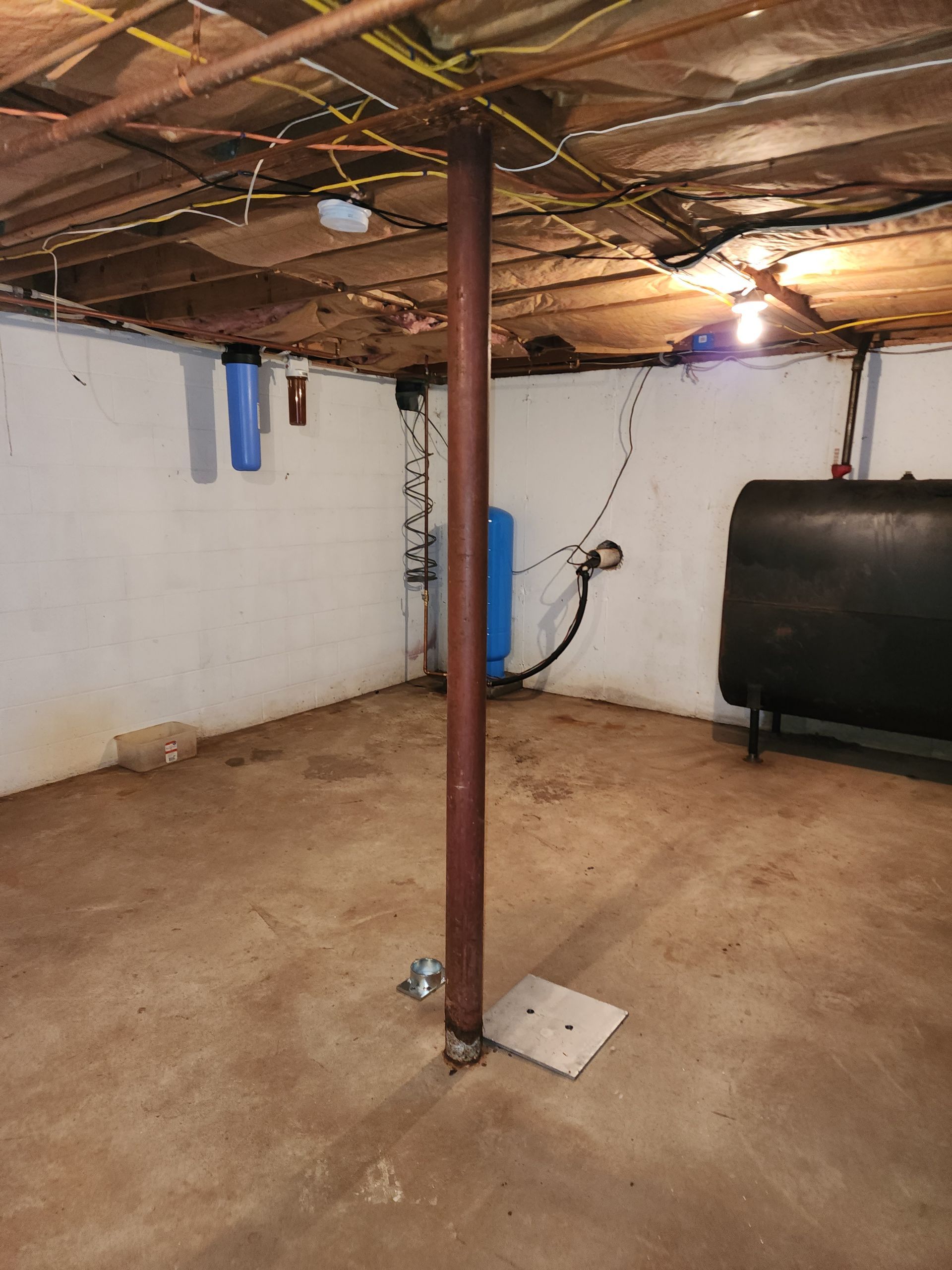 A basement interior featuring a central metal support column, a water filtration system, and a black storage tank.