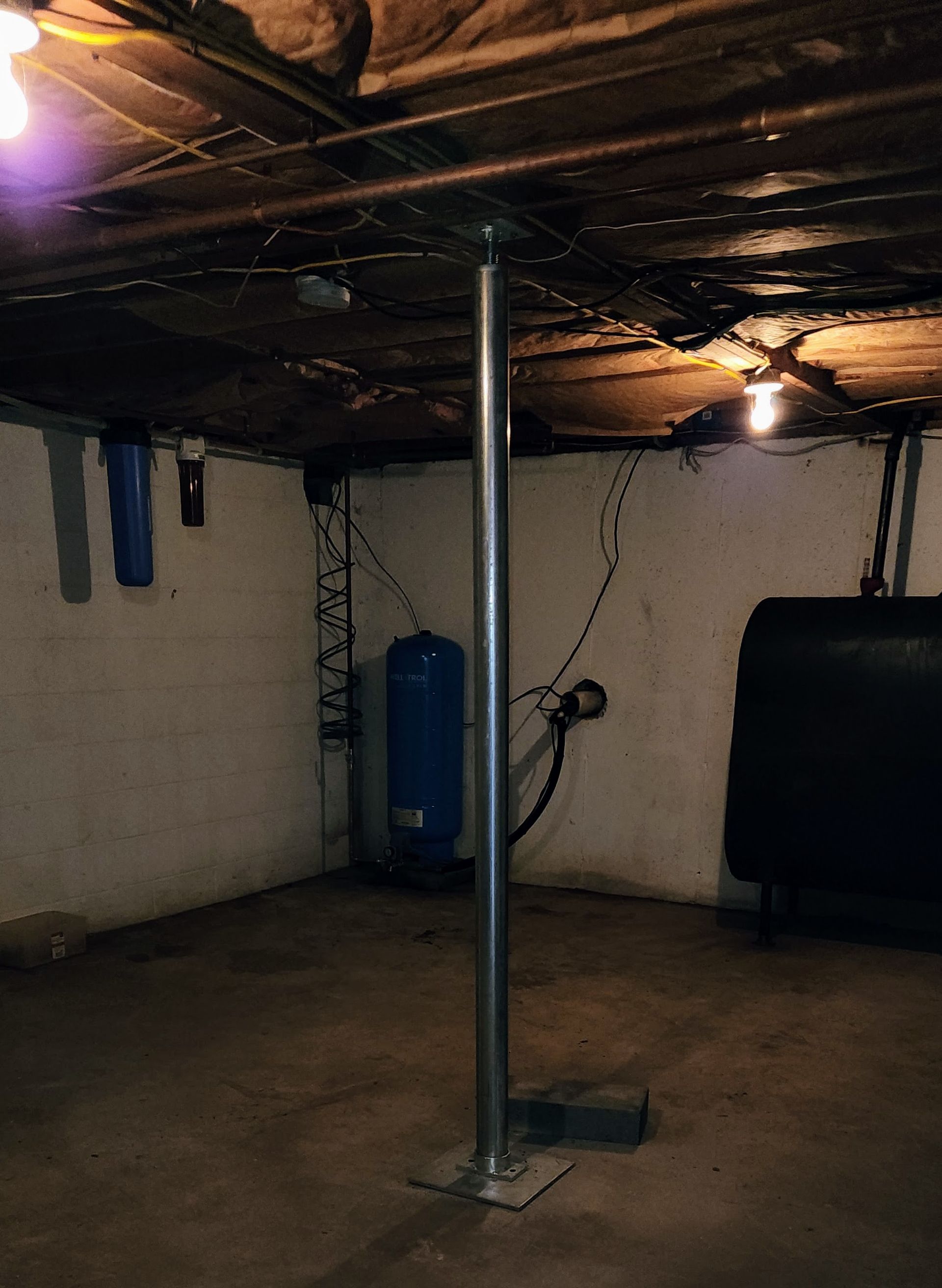 A steel support column stands in the center of a dim, unfinished concrete basement with utility equipment in the background.
