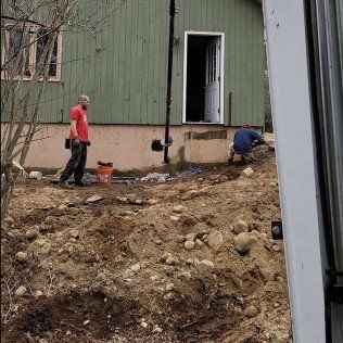 Two men working near a green house; one standing, the other squatting. Excavated dirt and rocks in foreground.