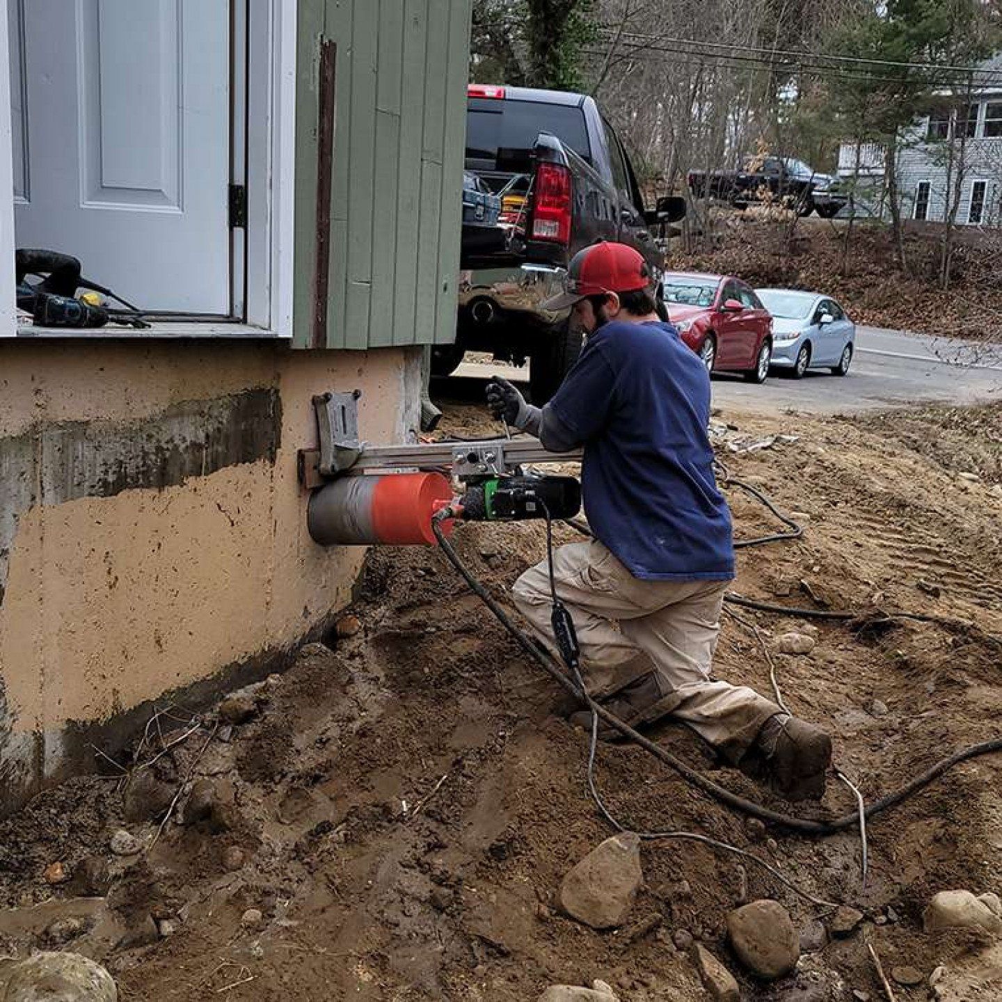 Man using a drill to repair a concrete foundation. Outdoors, dirt and a truck visible.
