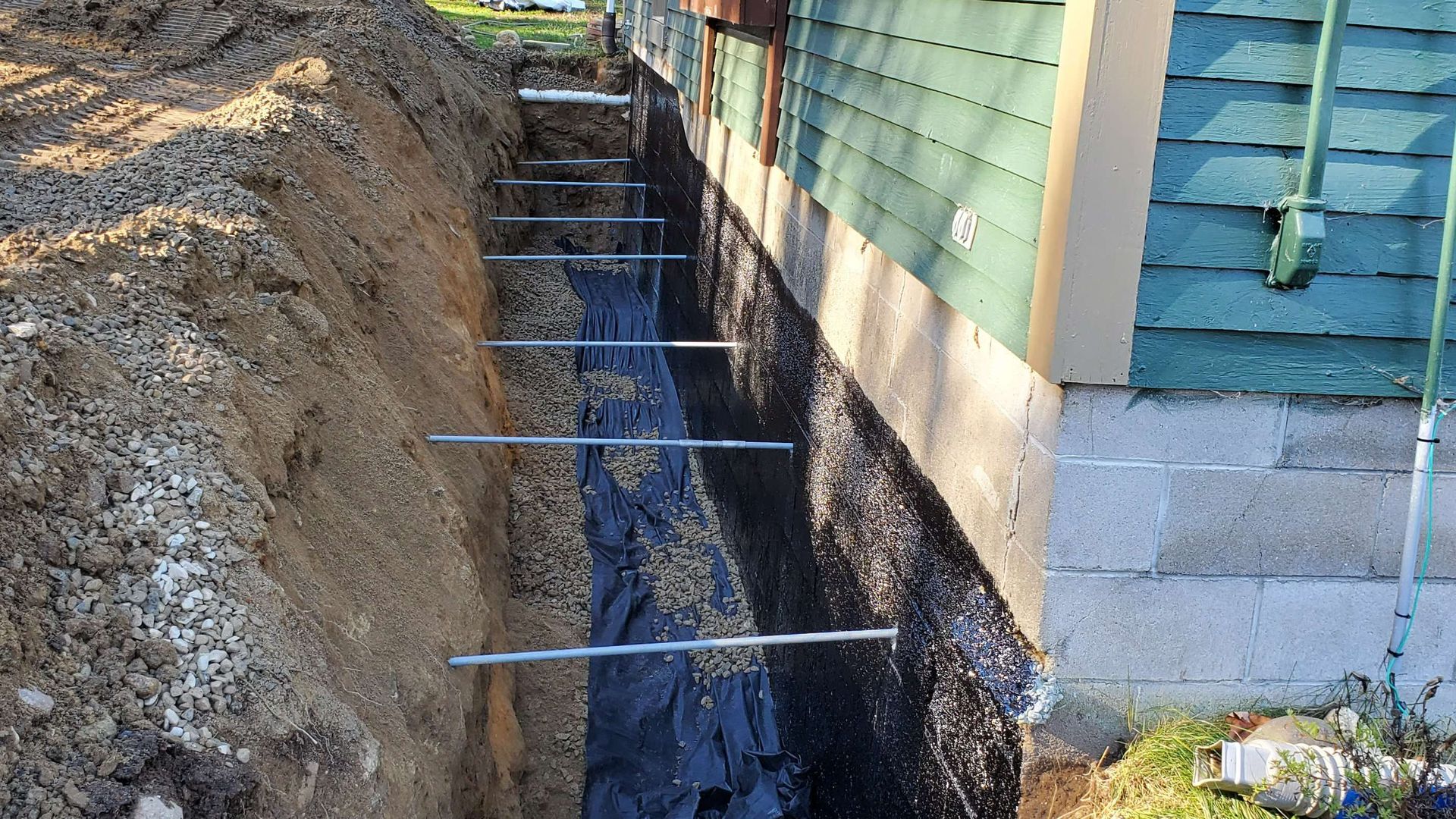 Trench dug beside a building, with metal tiebacks installed, and covered with black fabric, soil on left.