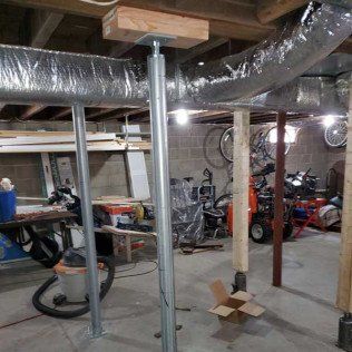 Basement with metal support jacks holding up ductwork; visible tools and bikes in the background.