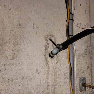 Concrete basement wall with a pipe and wires, showing water damage and a utility access point.