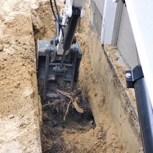 Mini excavator digging a trench near a building foundation, with exposed soil and roots.