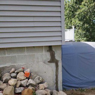 Corner of a house with gray siding, concrete block foundation, rocks, and a vertical water stain.
