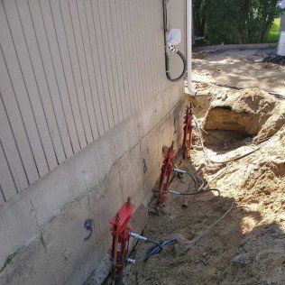 Foundation repair with hydraulic jacks; dirt trench along building, concrete foundation visible.