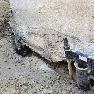 Construction workers excavating around a concrete wall, with tools and dirt visible.