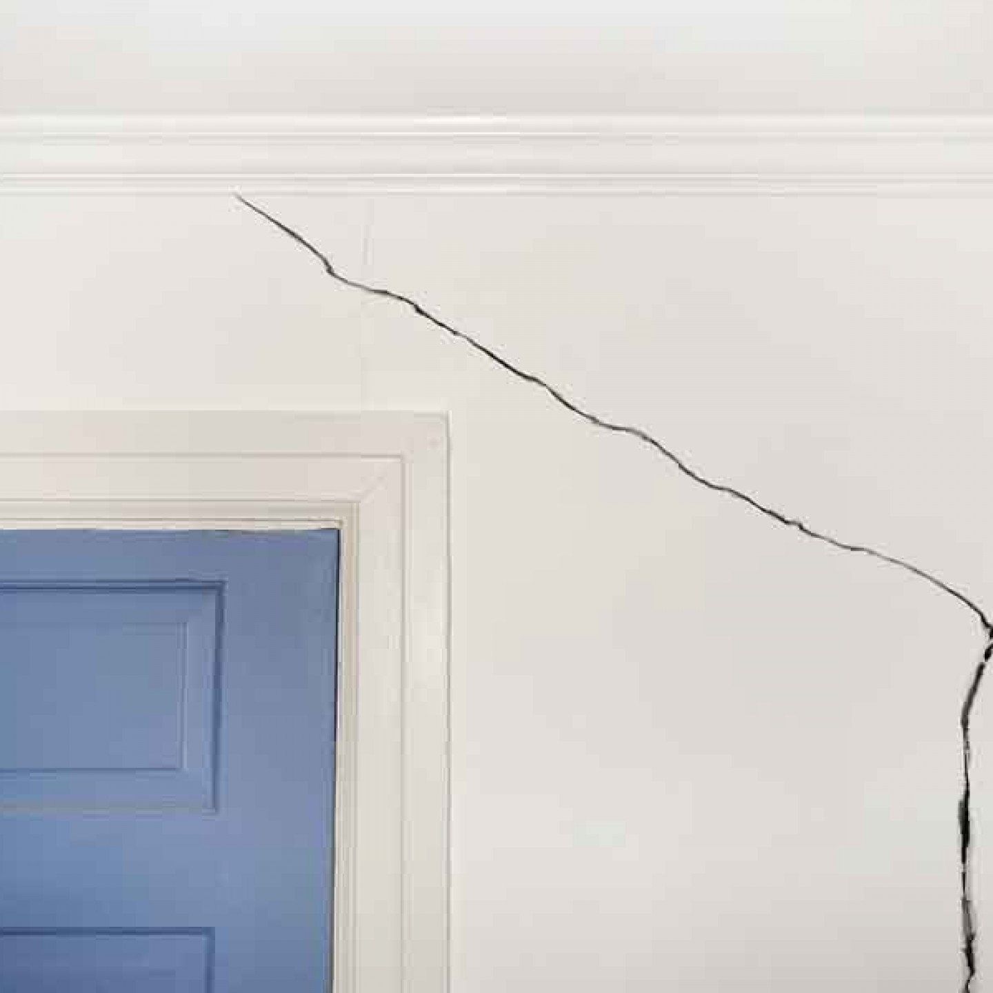 Crack in a white wall, near a blue door, extending from the ceiling.