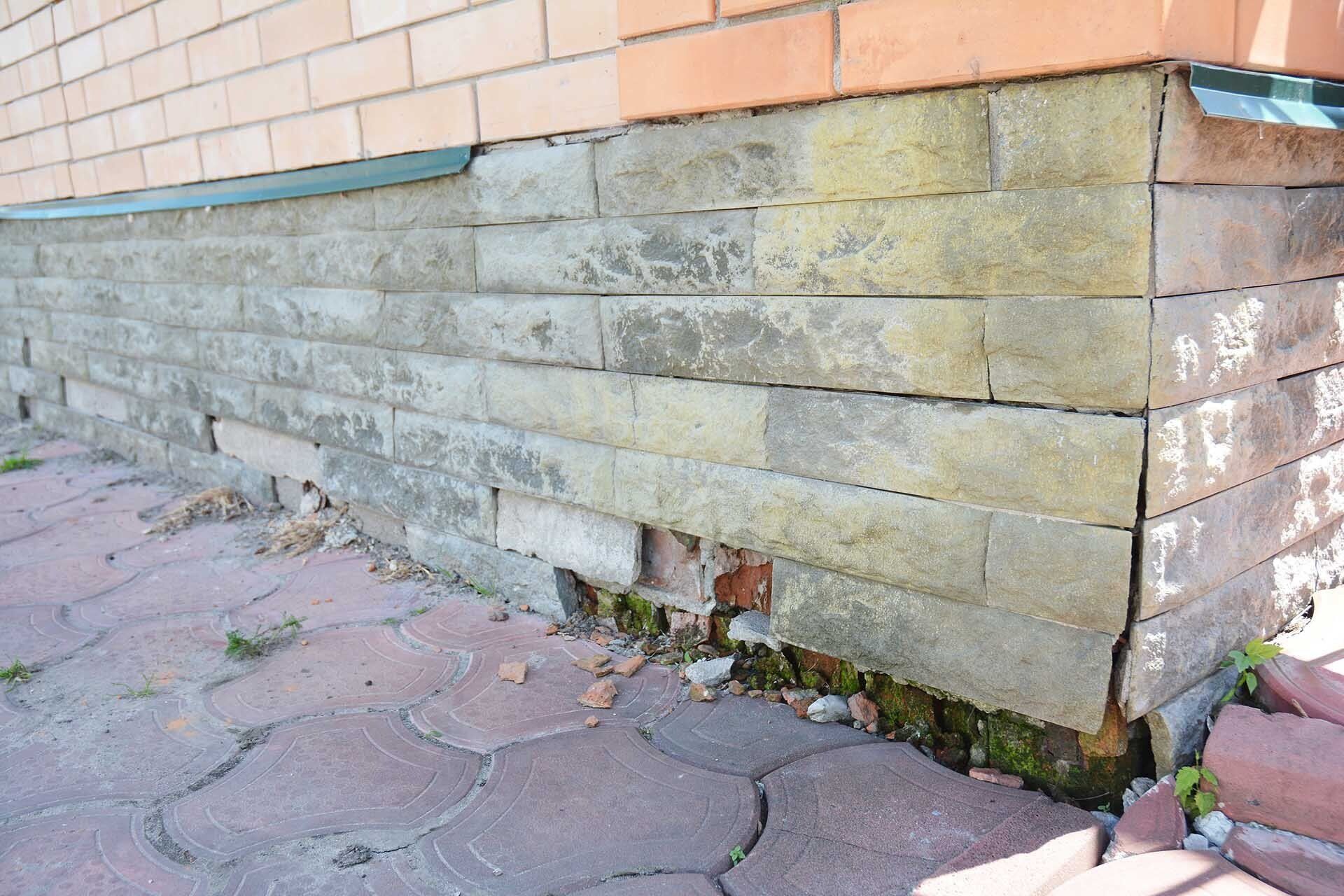 Brick foundation of a building with damaged bricks, moss, and cracked pavement.