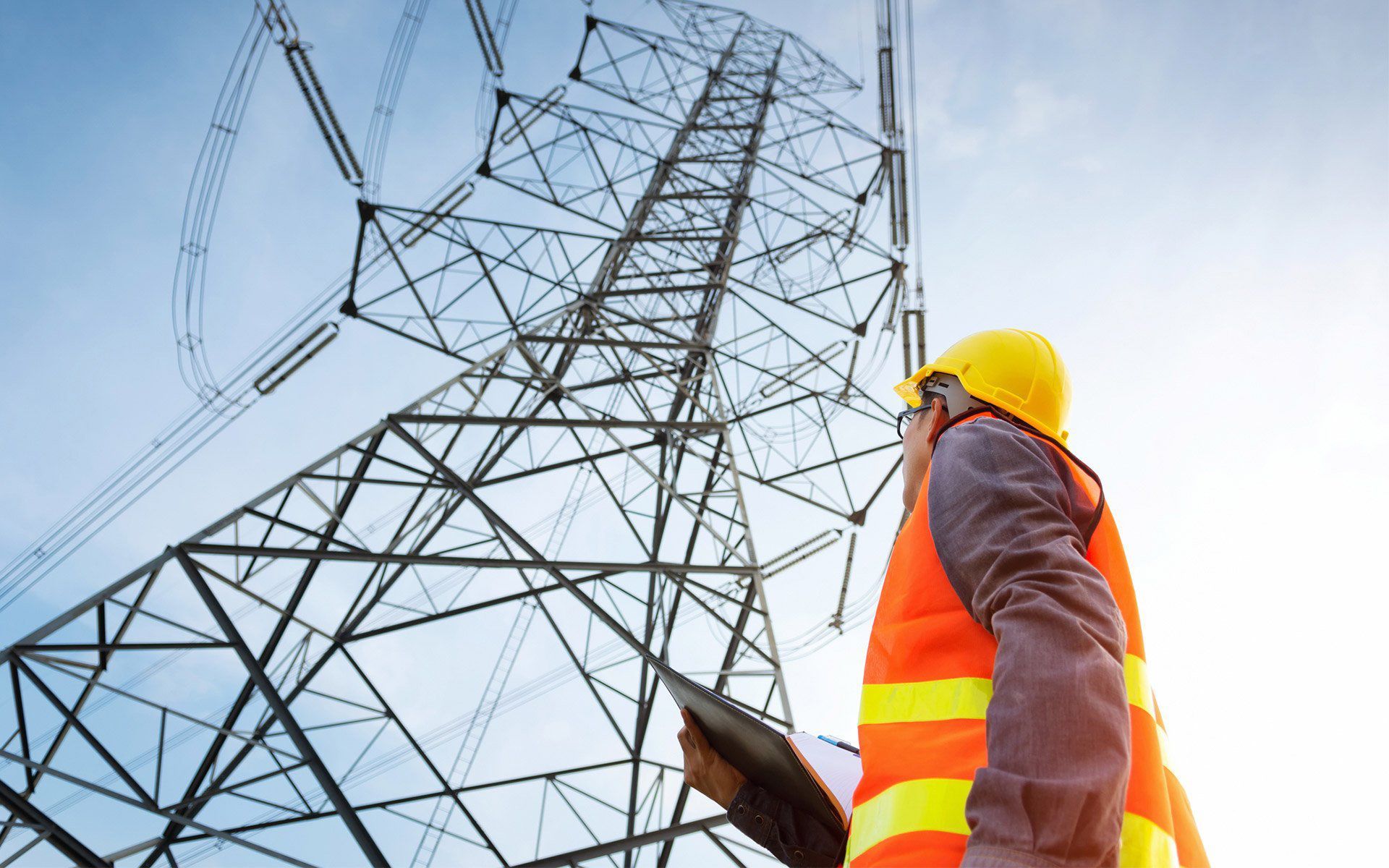 Engineer in safety vest and hard hat inspecting a large power pylon against a blue sky.