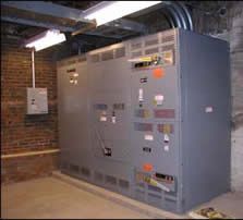 Gray electrical panel in a basement with brick walls, fluorescent lights, and exposed conduit.