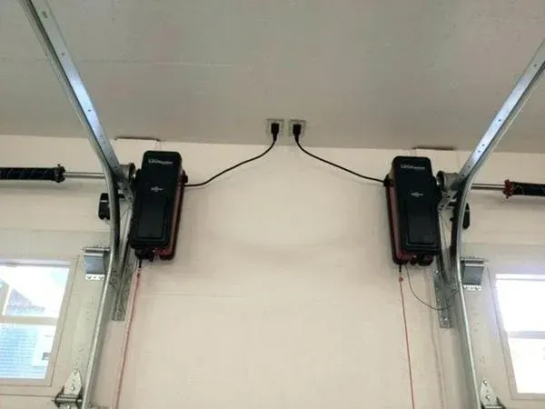 Two garage door openers mounted on a white wall, plugged into an electrical outlet.