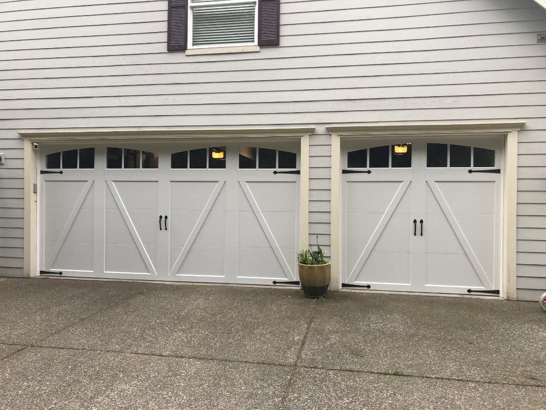 Two white garage doors with black hardware, arched top, and windows, set in a light gray house.