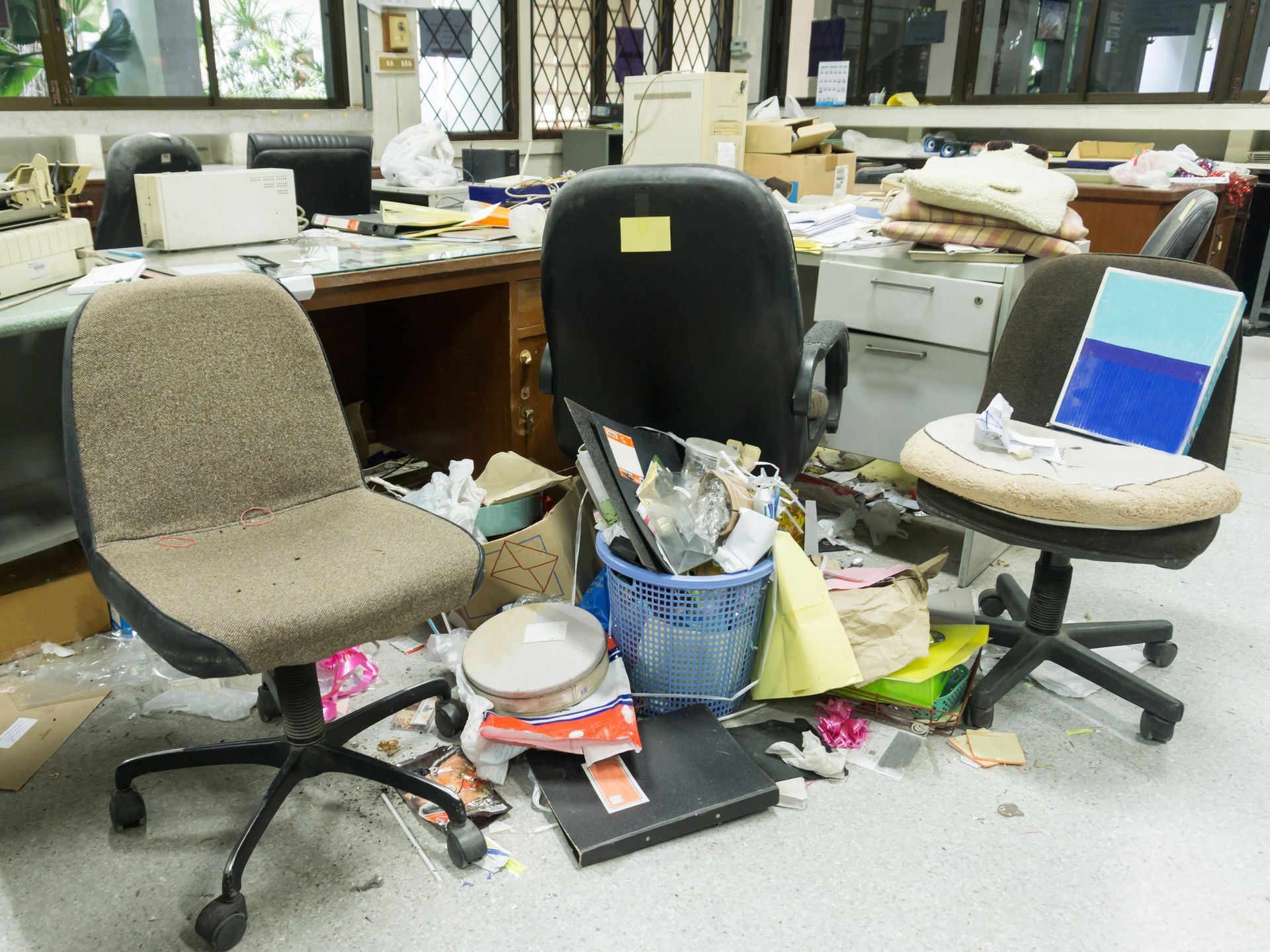 A messy office with two chairs , a desk , and a trash can.