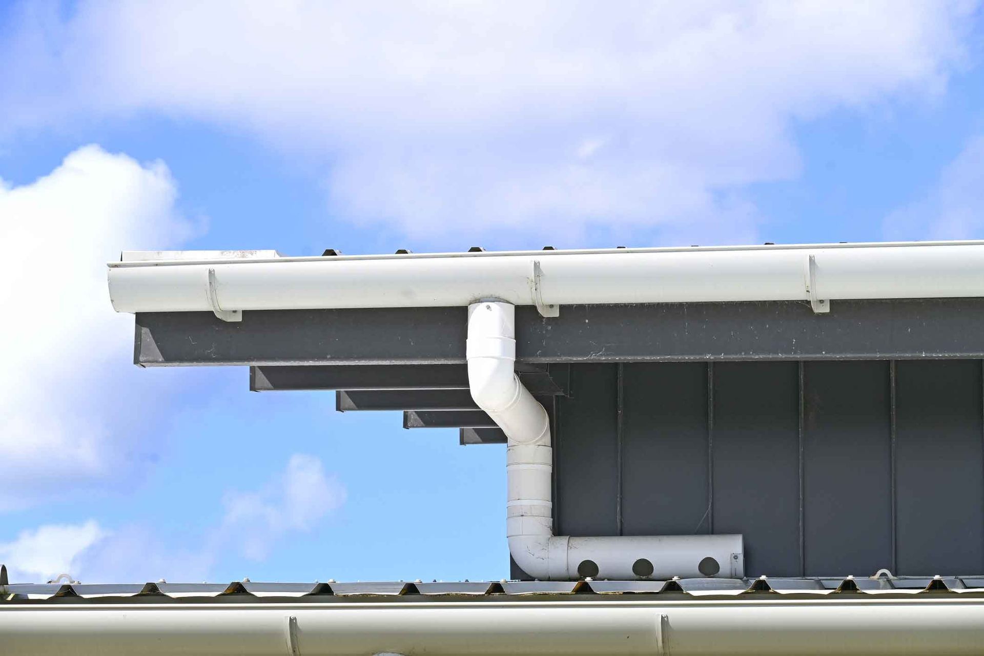 A white gutter and downpipe on the roof of a house with a blue sky in the background.