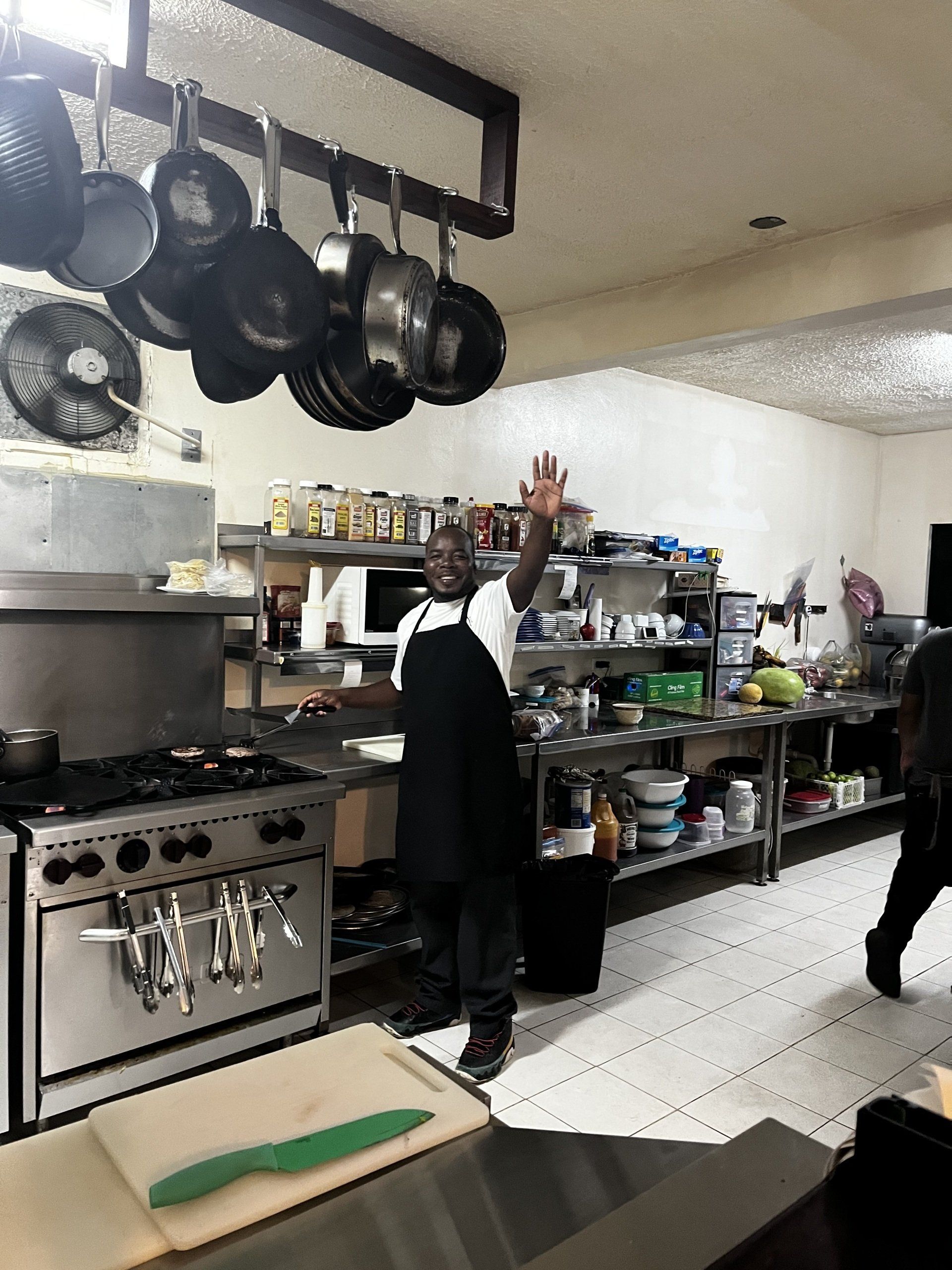 A man in an apron is standing in a kitchen with pots and pans hanging from the ceiling