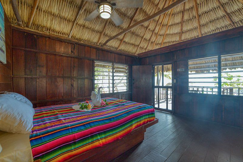 A bedroom with a bed , ceiling fan , and thatched roof.