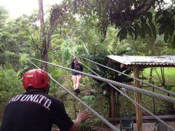 A man in a black shirt with the word tunito on the back is watching a woman on a zip line.