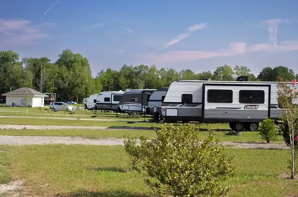 Campground with RVs parked on grass, small building, cars, trees, and blue sky.
