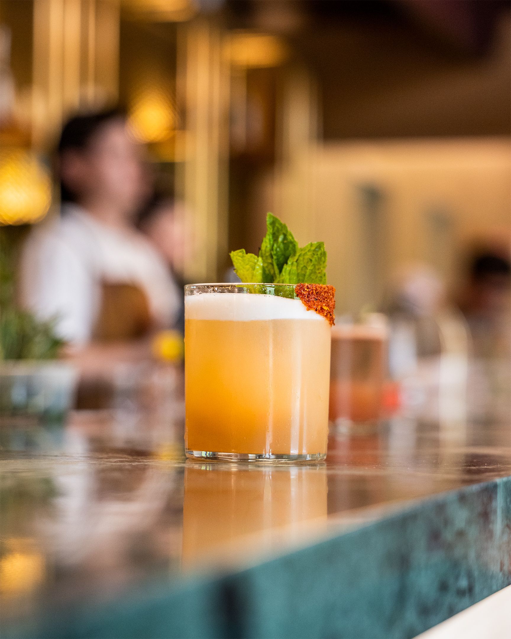 Cocktail on a bar, garnished with mint and a citrus slice; blurry background of a bar scene.