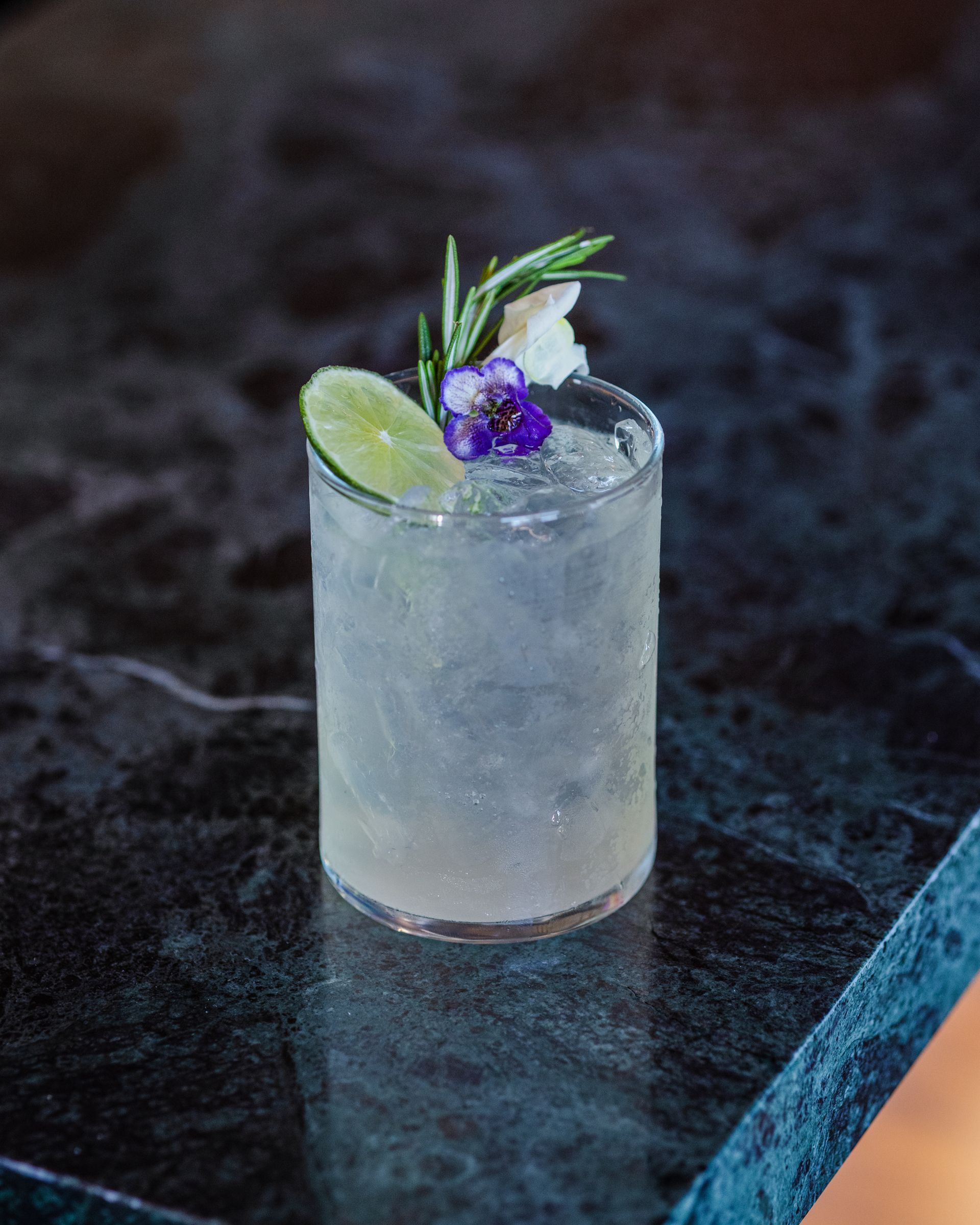 Cocktail in a glass garnished with lime, flower, rosemary, on a dark countertop.