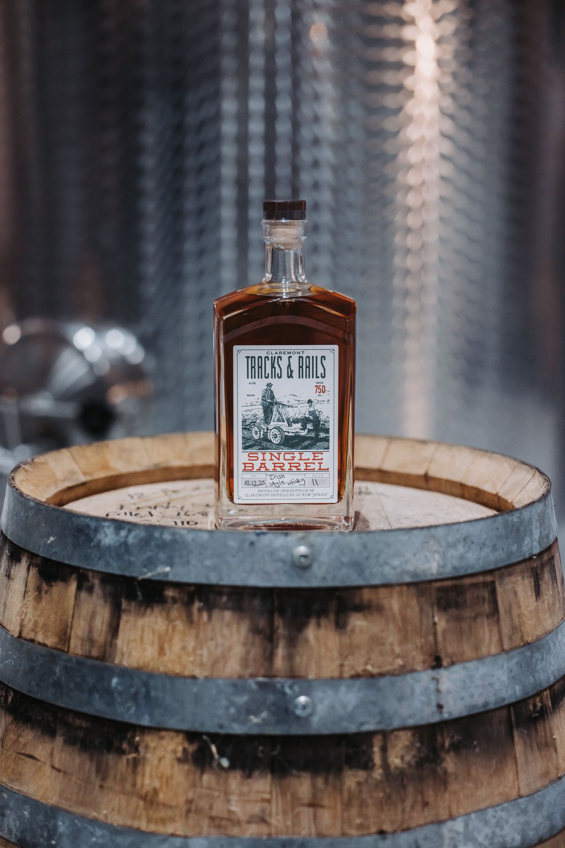 Bottle of amber liquid with label on wooden barrel, in front of a metal tank.