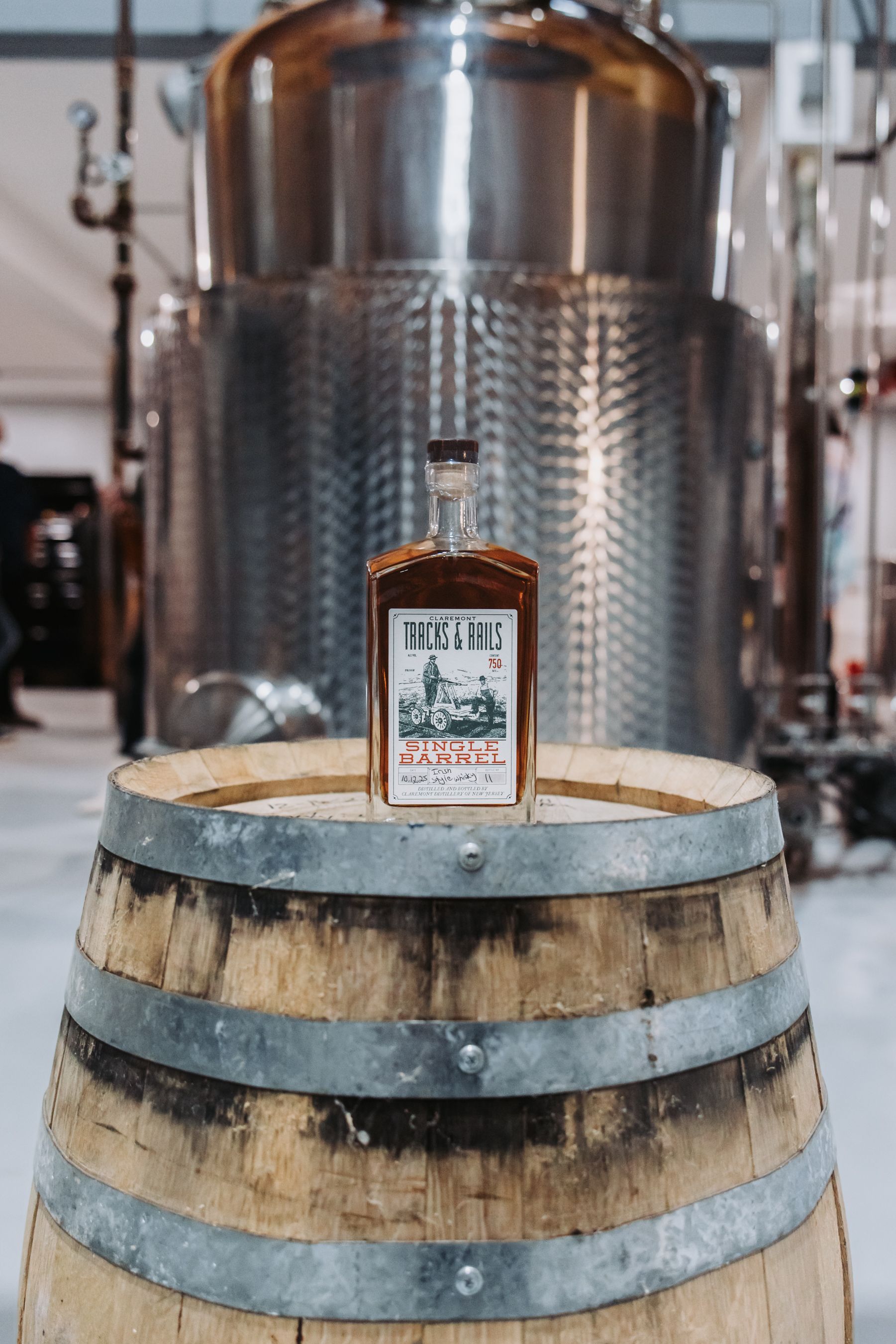 Bottle of whiskey on a wooden barrel in a distillery with stainless steel tanks.
