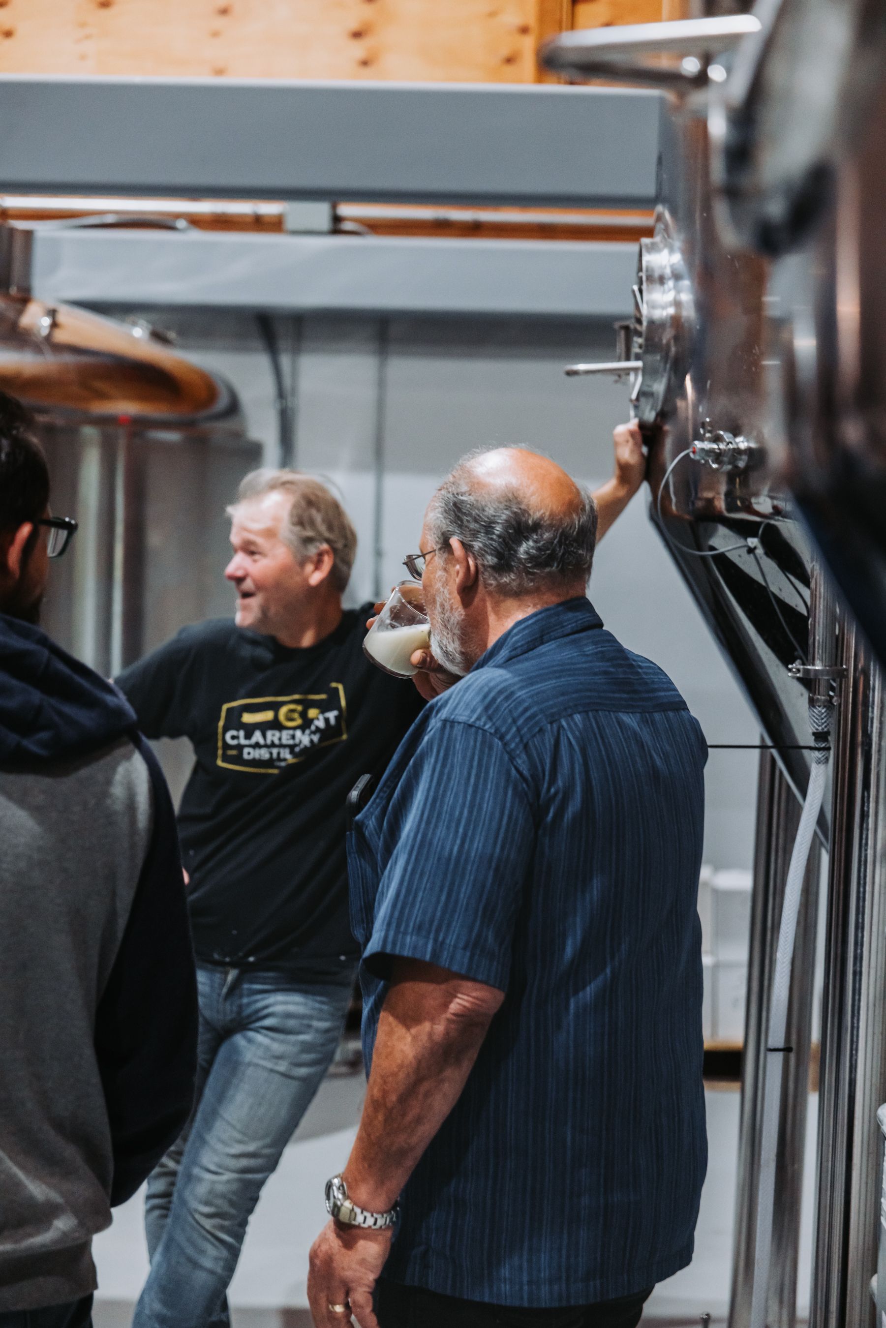 Three men in a brewery, inspecting equipment; one smiles, others look on.