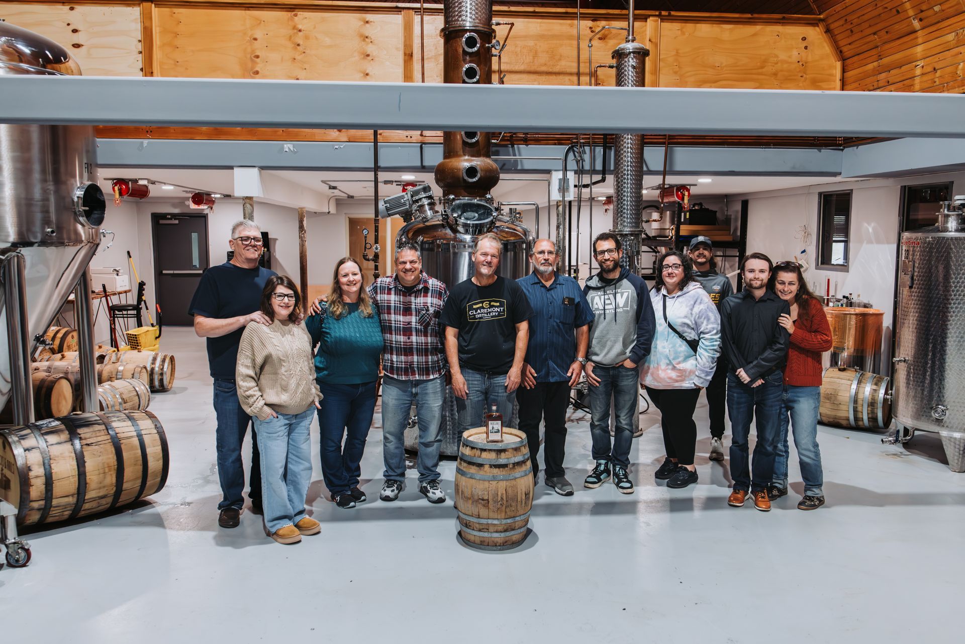 Group of people posing in distillery with equipment and barrels.