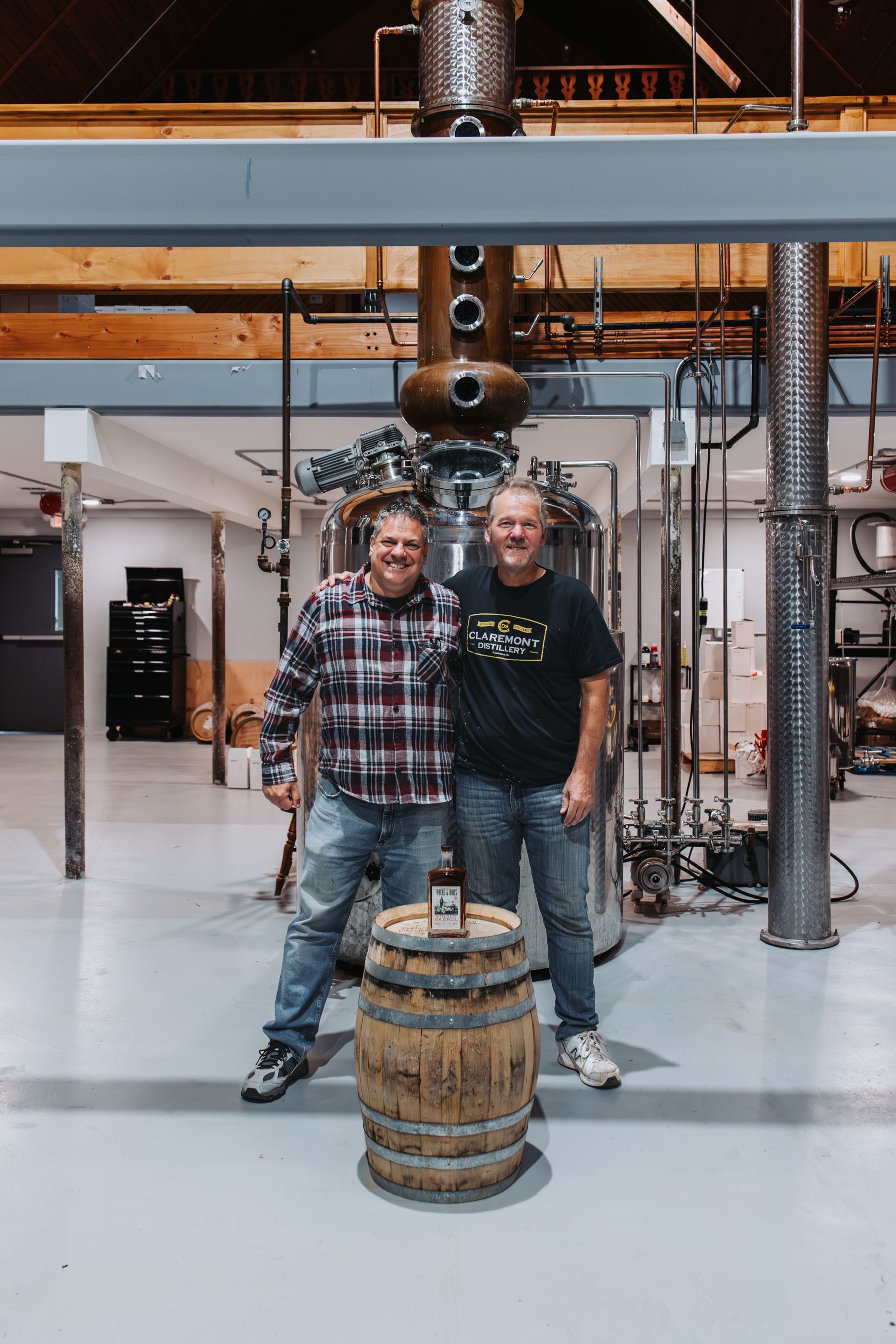 Two men stand near a distillery barrel. Behind them is stainless steel equipment in a large room.