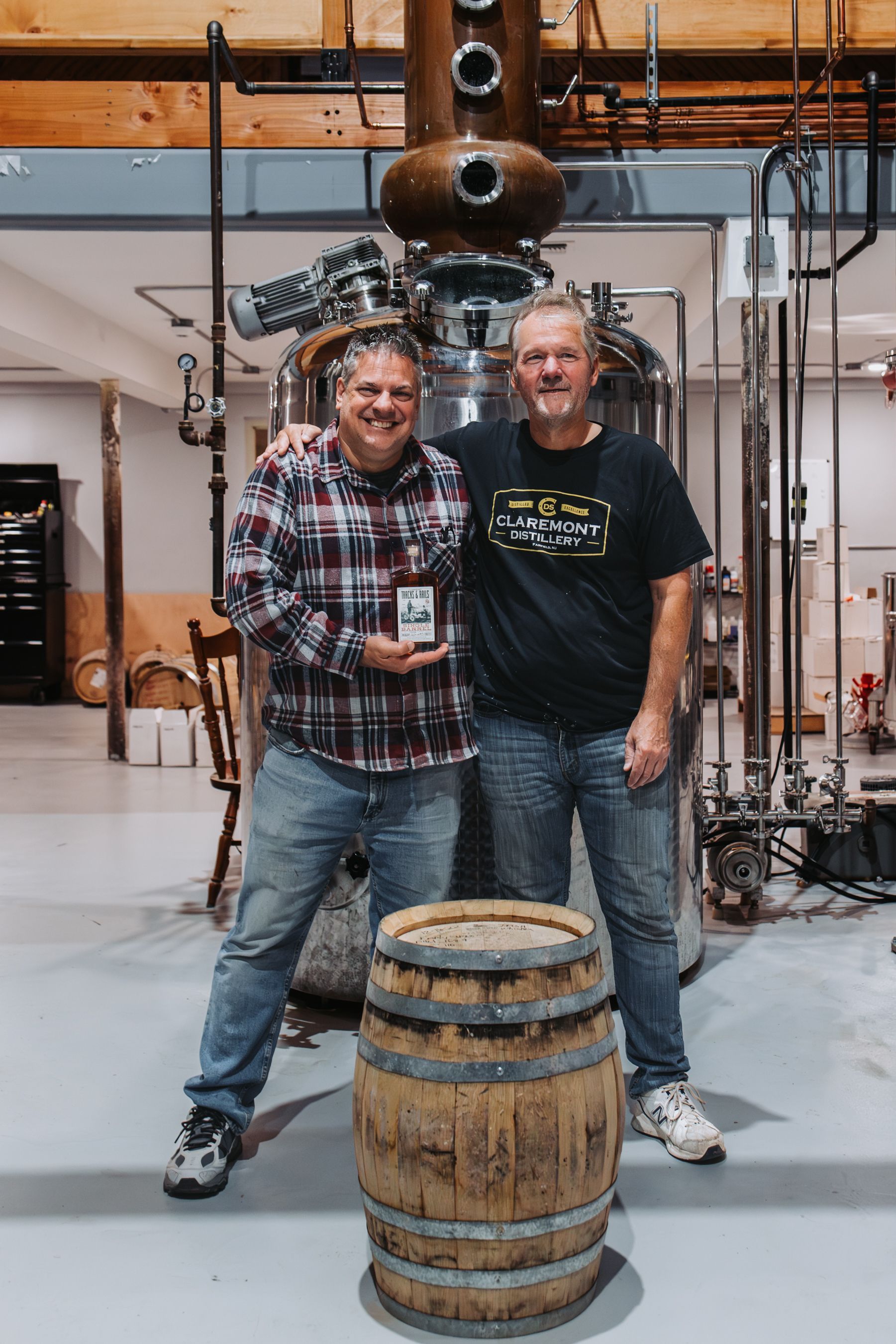 Two men stand in a distillery. One holds an award, while the other has an arm around his shoulders, in front of an aging barrel.
