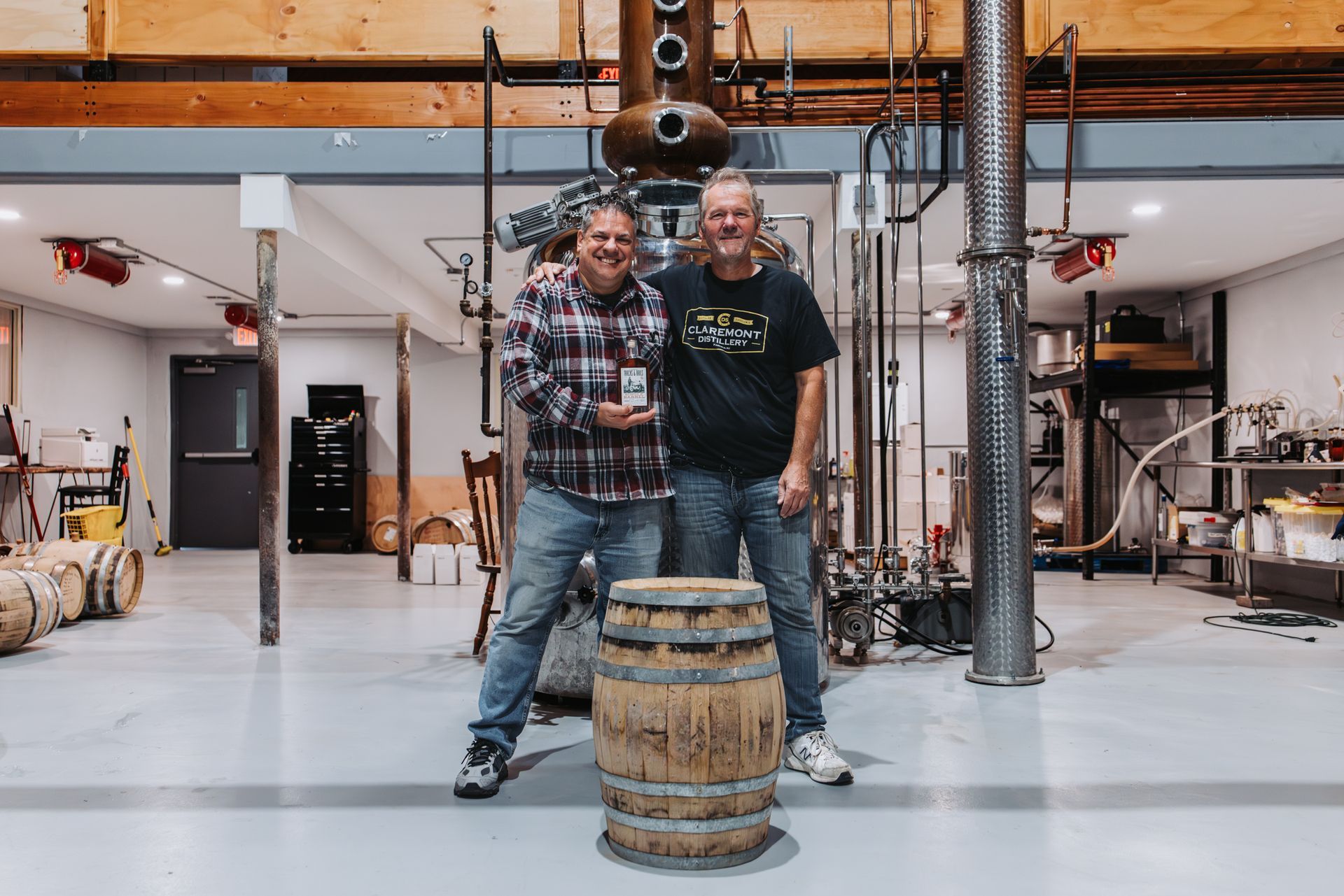 Two men pose with a liquor bottle in a distillery, barrel in front. One holds the bottle.