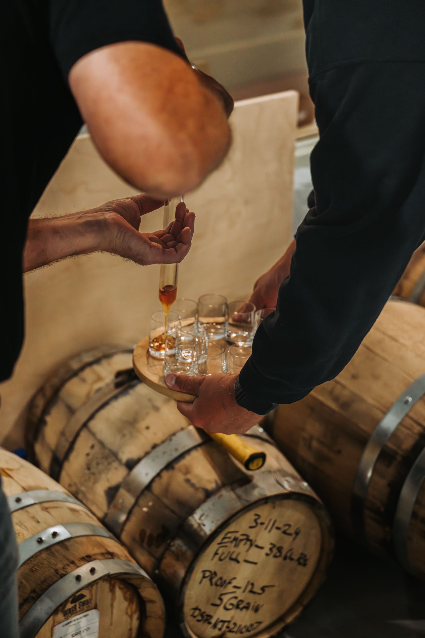 People pouring liquid into small glasses on a tray, near barrels.