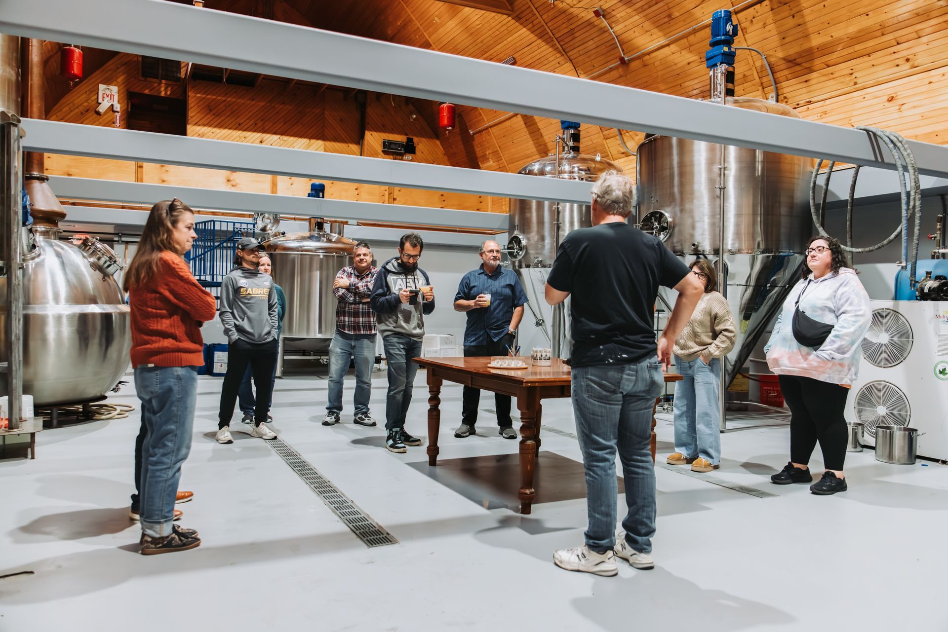 Group of people on a brewery tour, listening to a presentation in front of stainless steel tanks.
