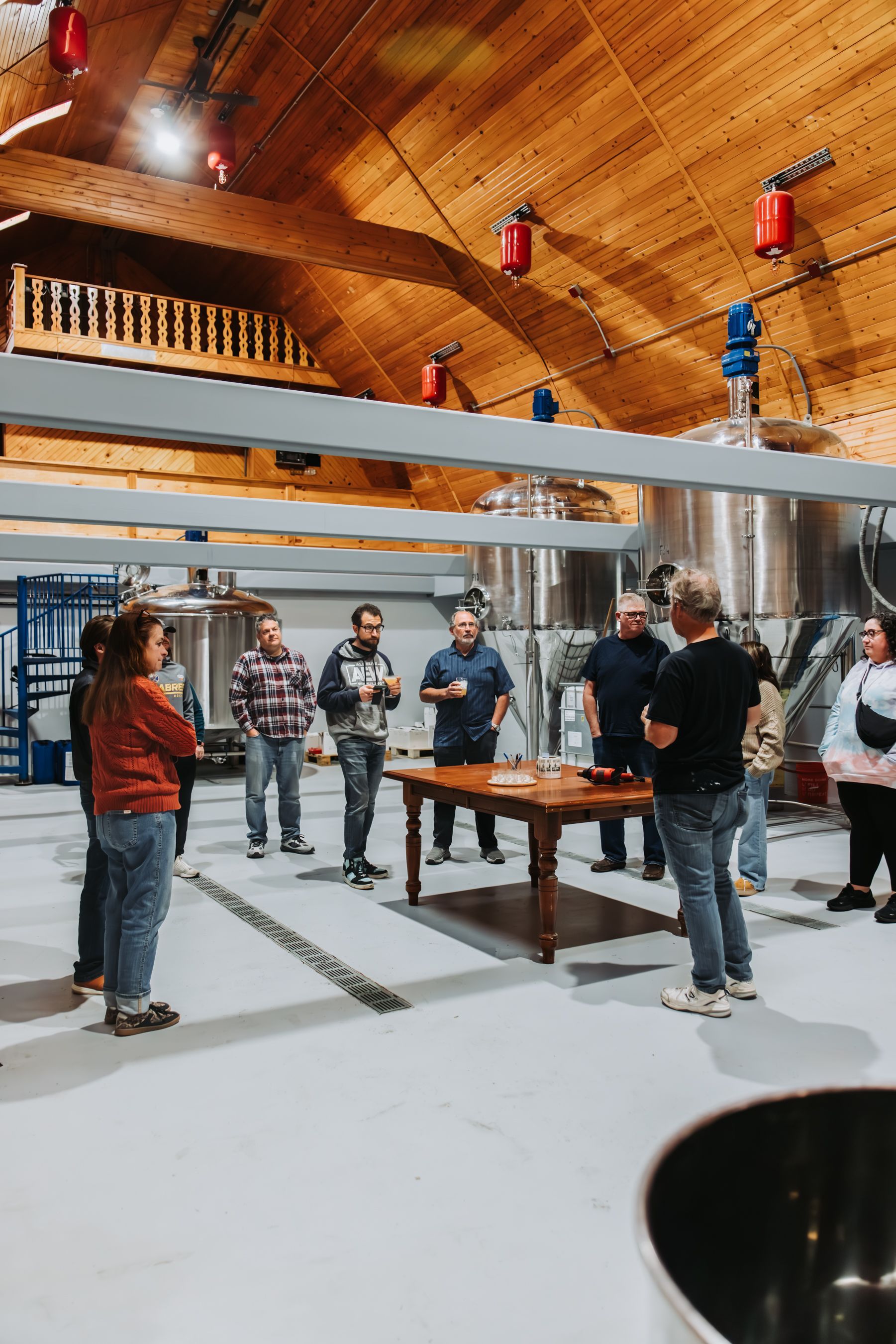 Group of people at a brewery tasting. They are standing around a table with drinks, with large silver tanks in background.