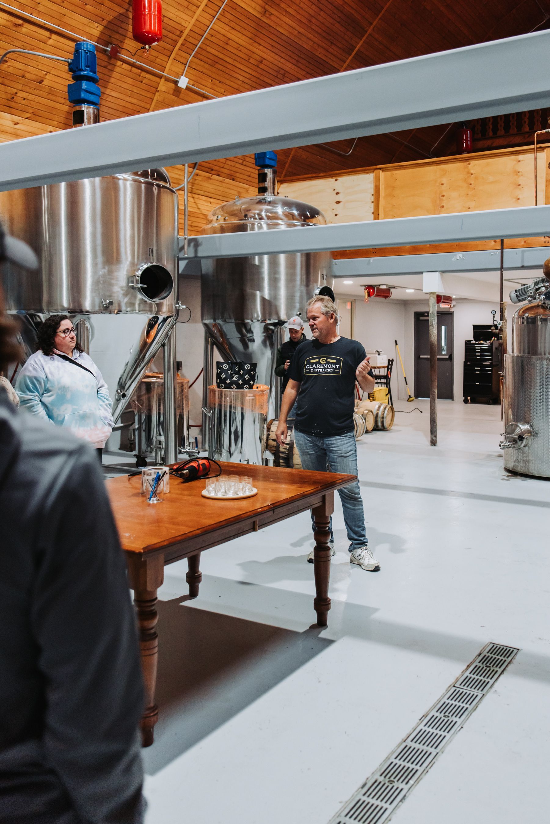 A man giving a tour in a brewery; people stand around a wooden table in a large, industrial space.