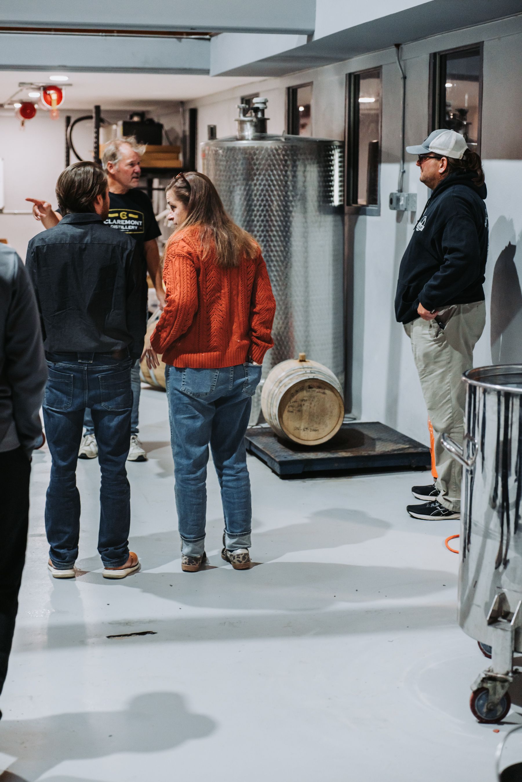 People in a winery, examining equipment. A woman in orange jacket and man in denim stand in front of a stainless steel tank.