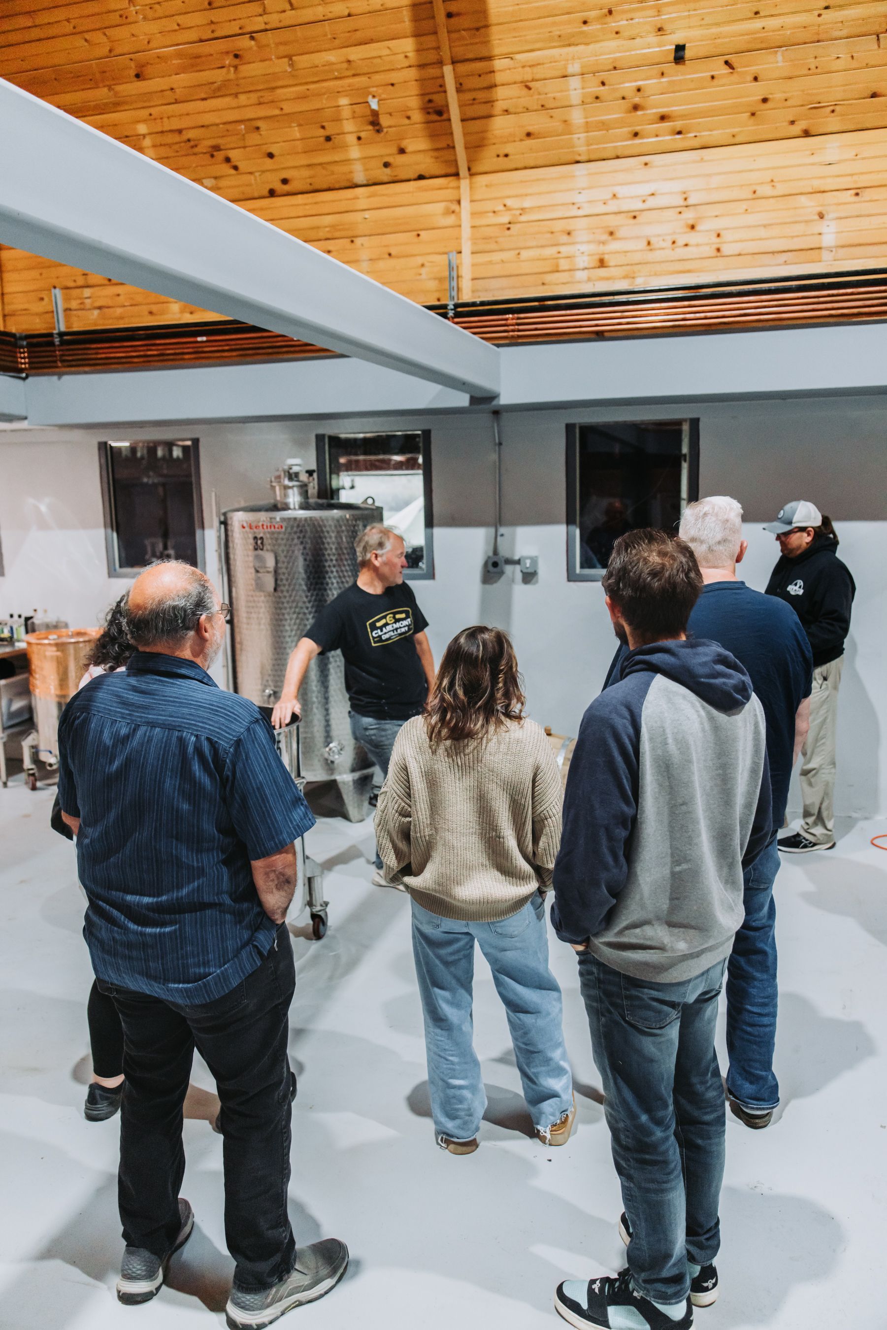 Group of people in a workshop, looking at a machine. Wooden ceiling, gray walls, and bright lighting.