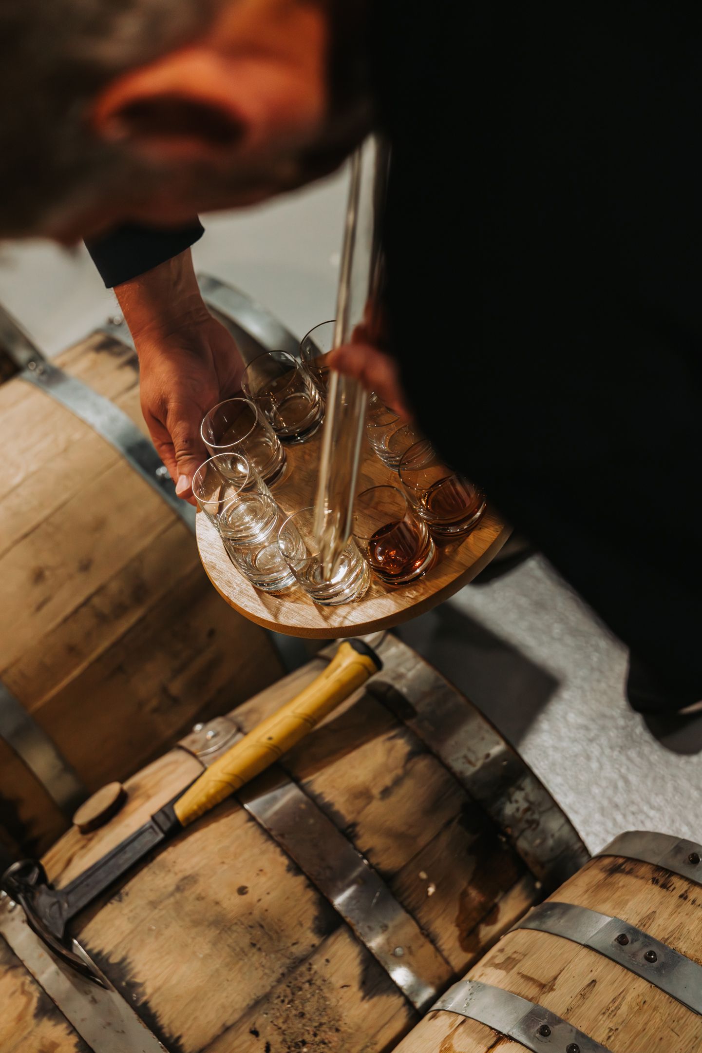 Person pouring liquid from a tube into small glasses on a wooden tray, near wooden barrels.