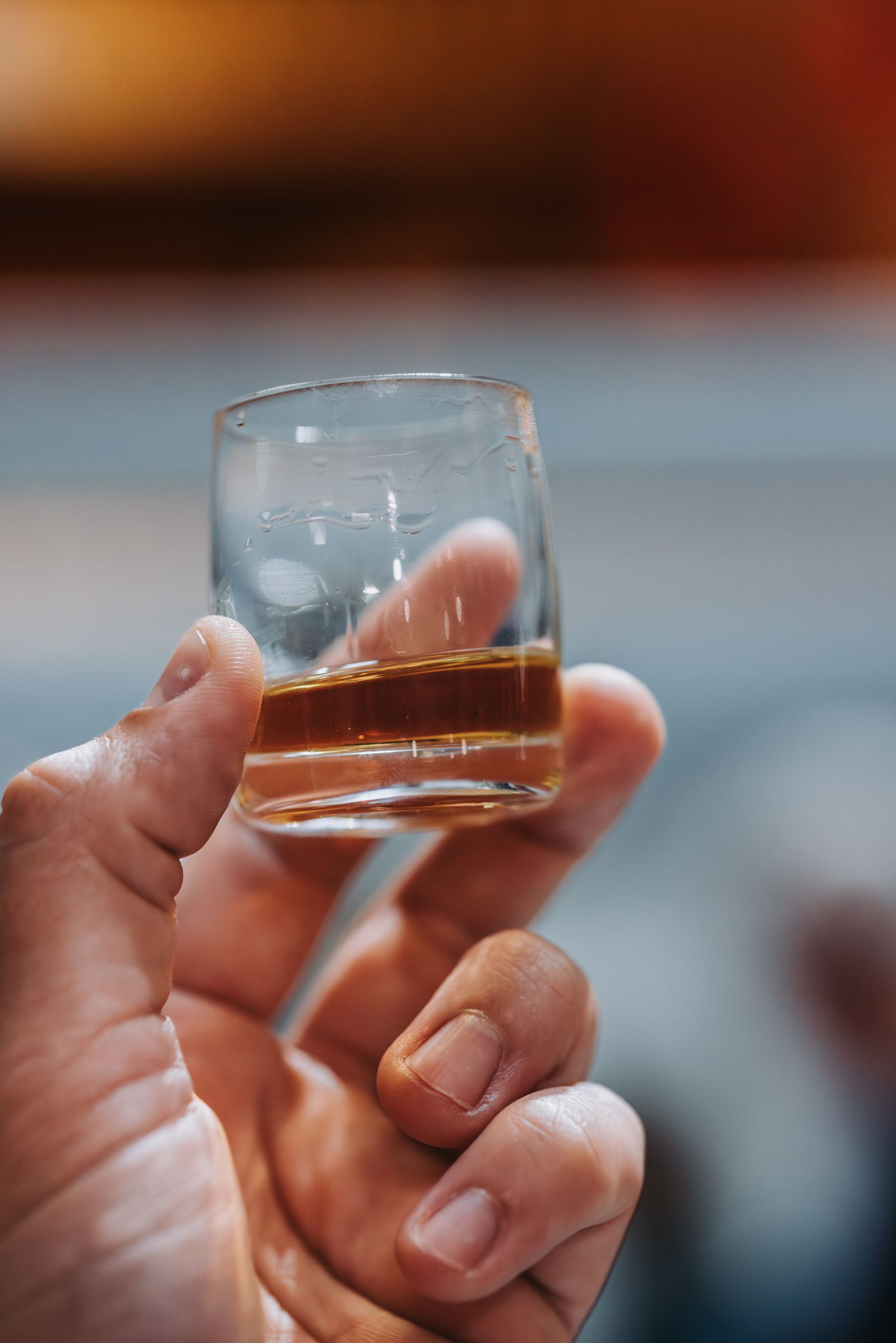Hand holding a small glass of amber-colored liquid; blurred background.