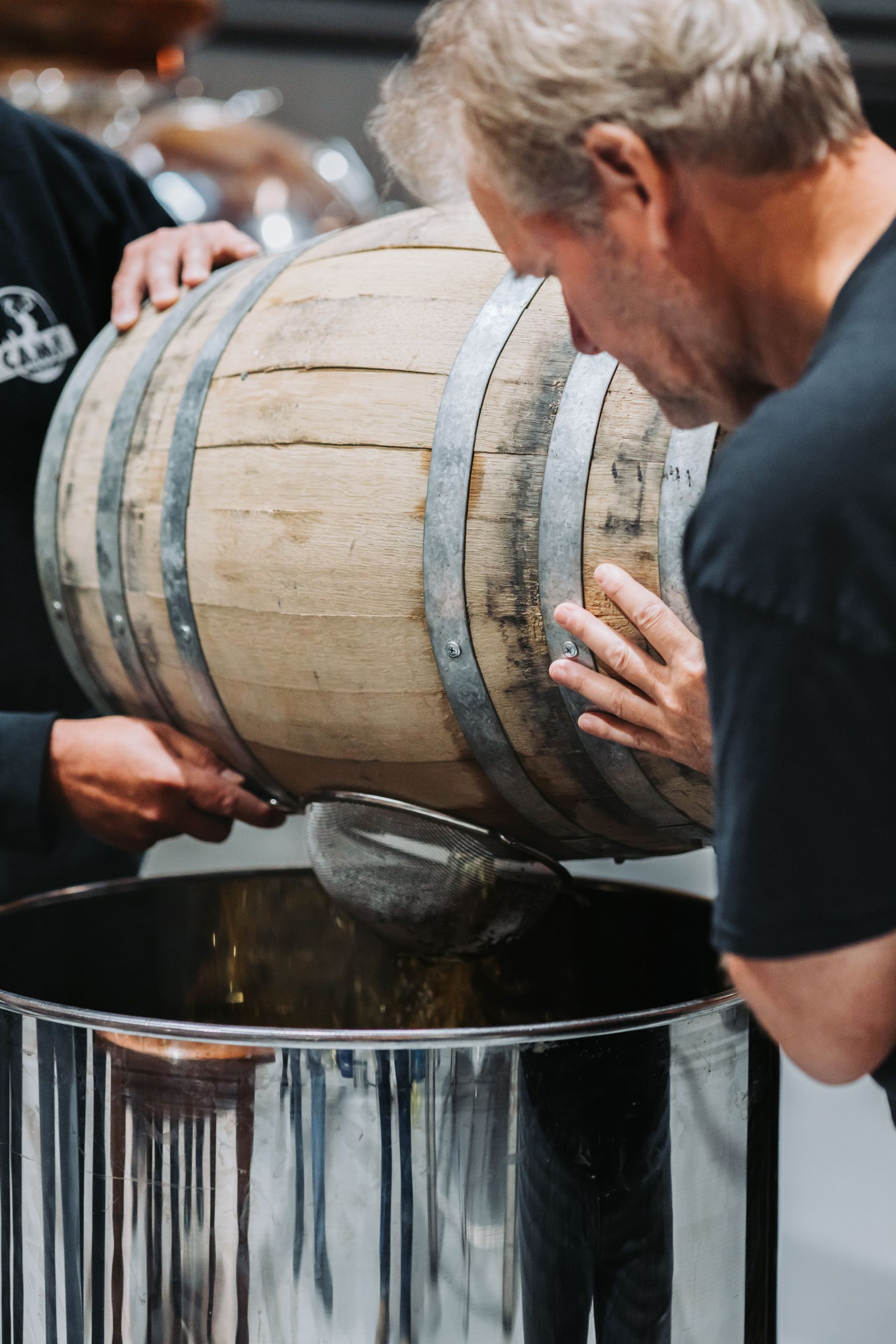 Two people pouring liquid from a wooden barrel into a stainless steel container. One man is smelling the barrel.