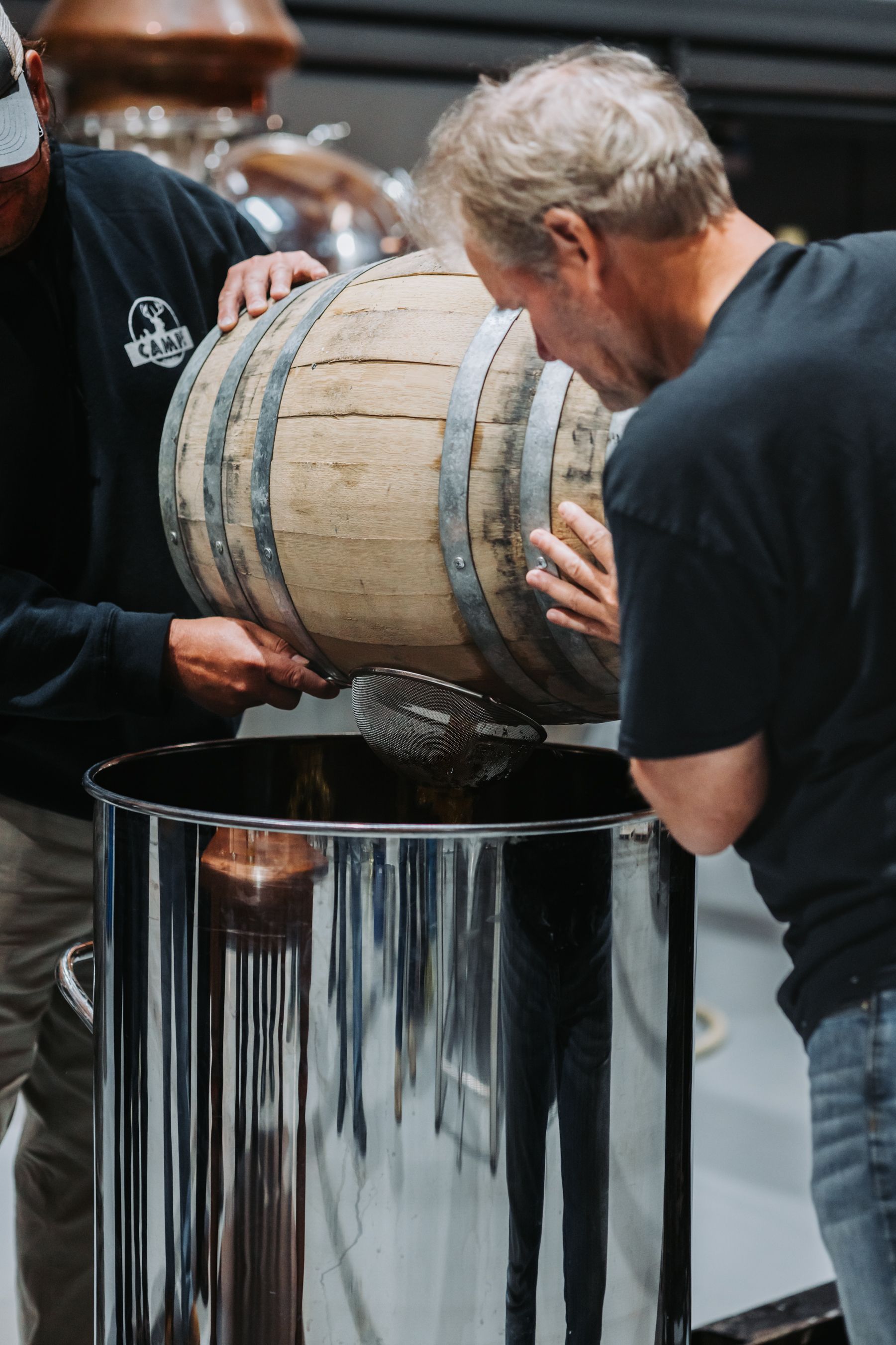 Two people pouring liquid from a wooden barrel into a large metal container in a distillery.