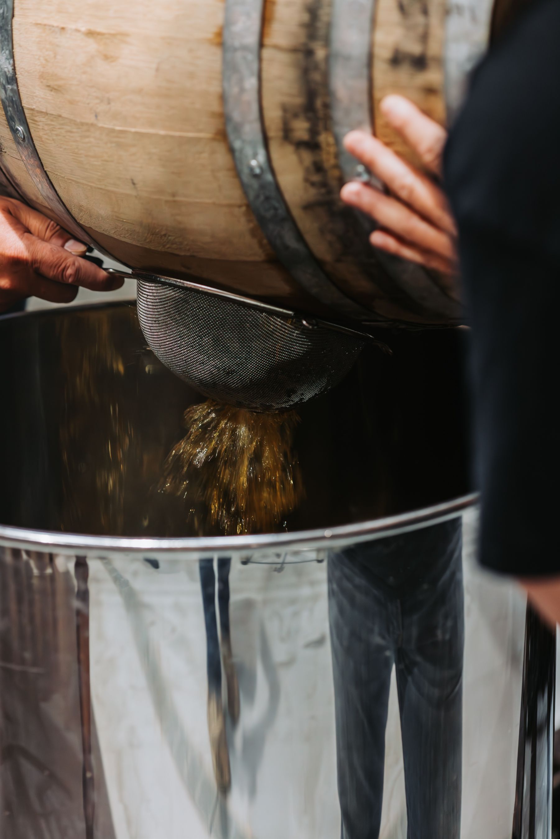 Liquid draining from a wooden barrel through a mesh strainer into a large stainless steel container.