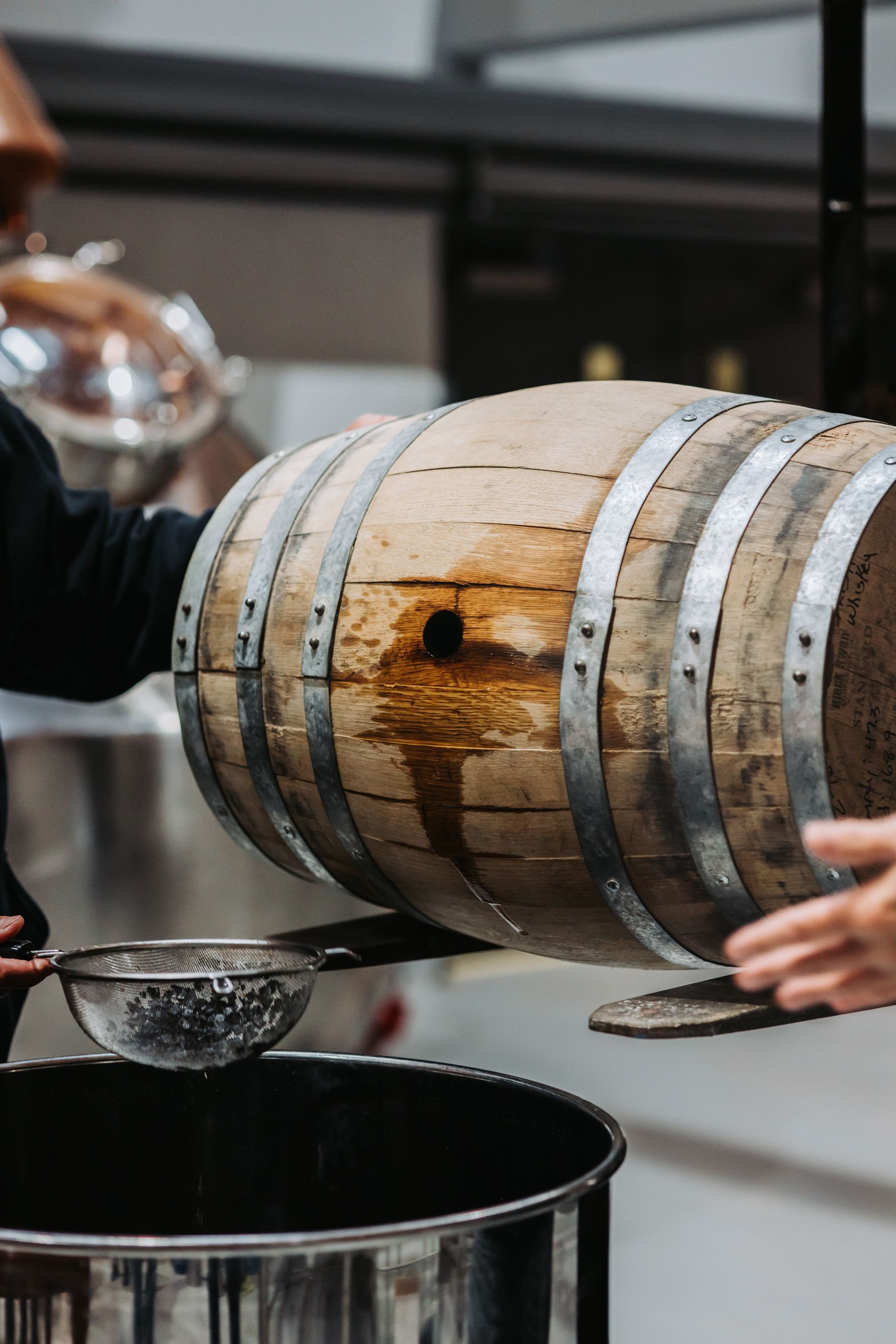 A barrel being tipped over, pouring liquid through a strainer into a metal container indoors.
