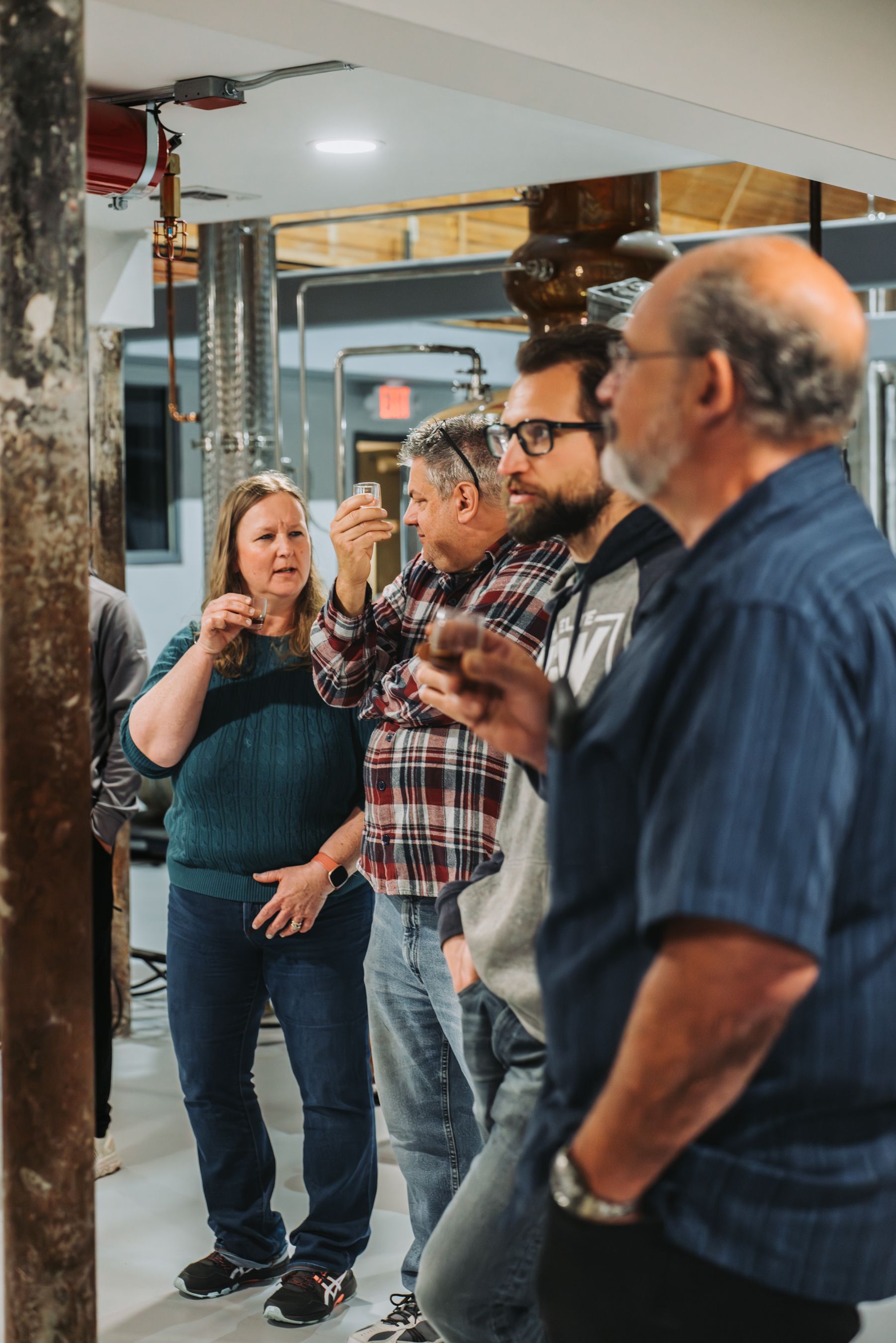 People tasting something in a dimly lit industrial space. Some hold glasses. One woman gestures while speaking.